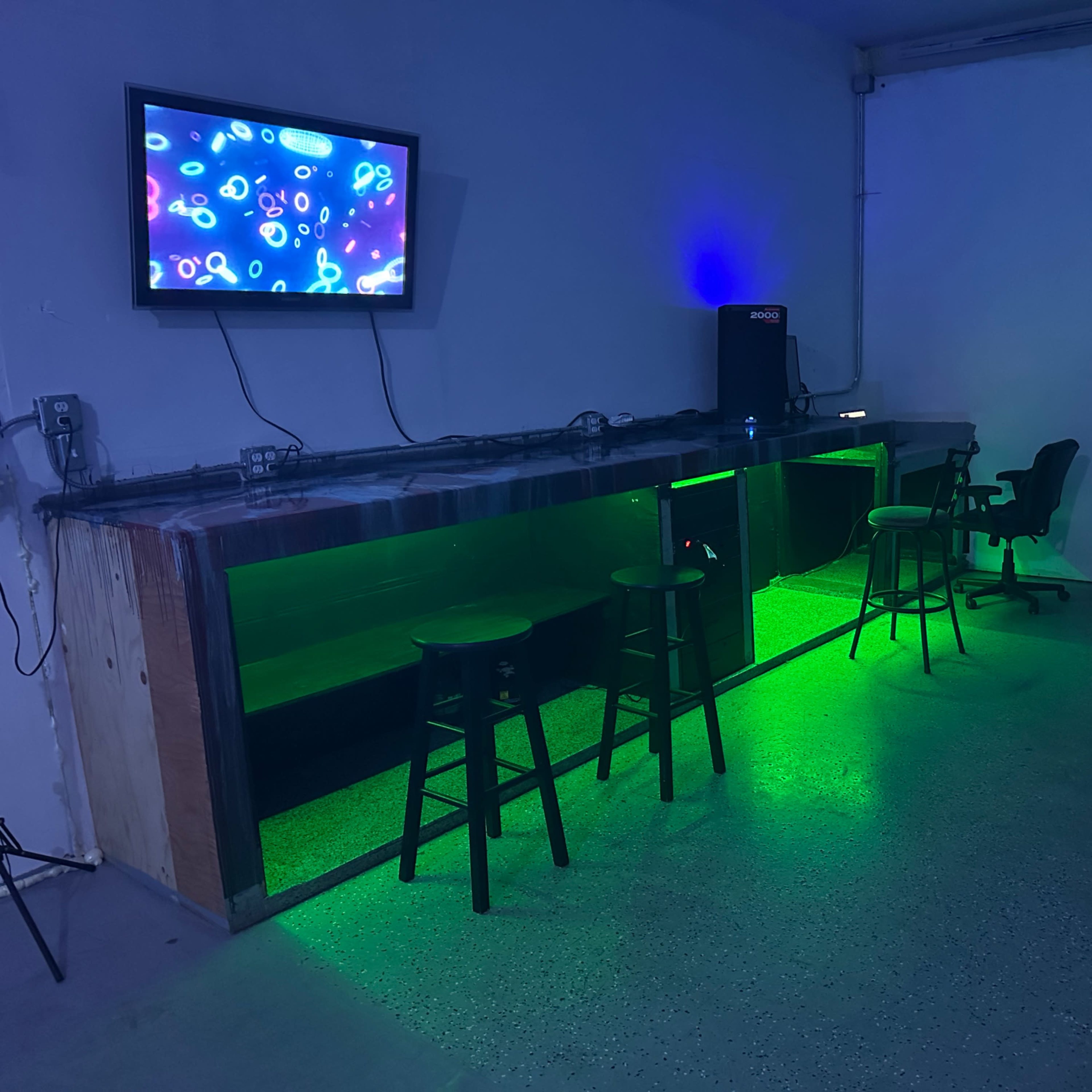 A dimly lit room featuring a bar with green lighting, several stools, a large screen on the wall, and a table with chairs.