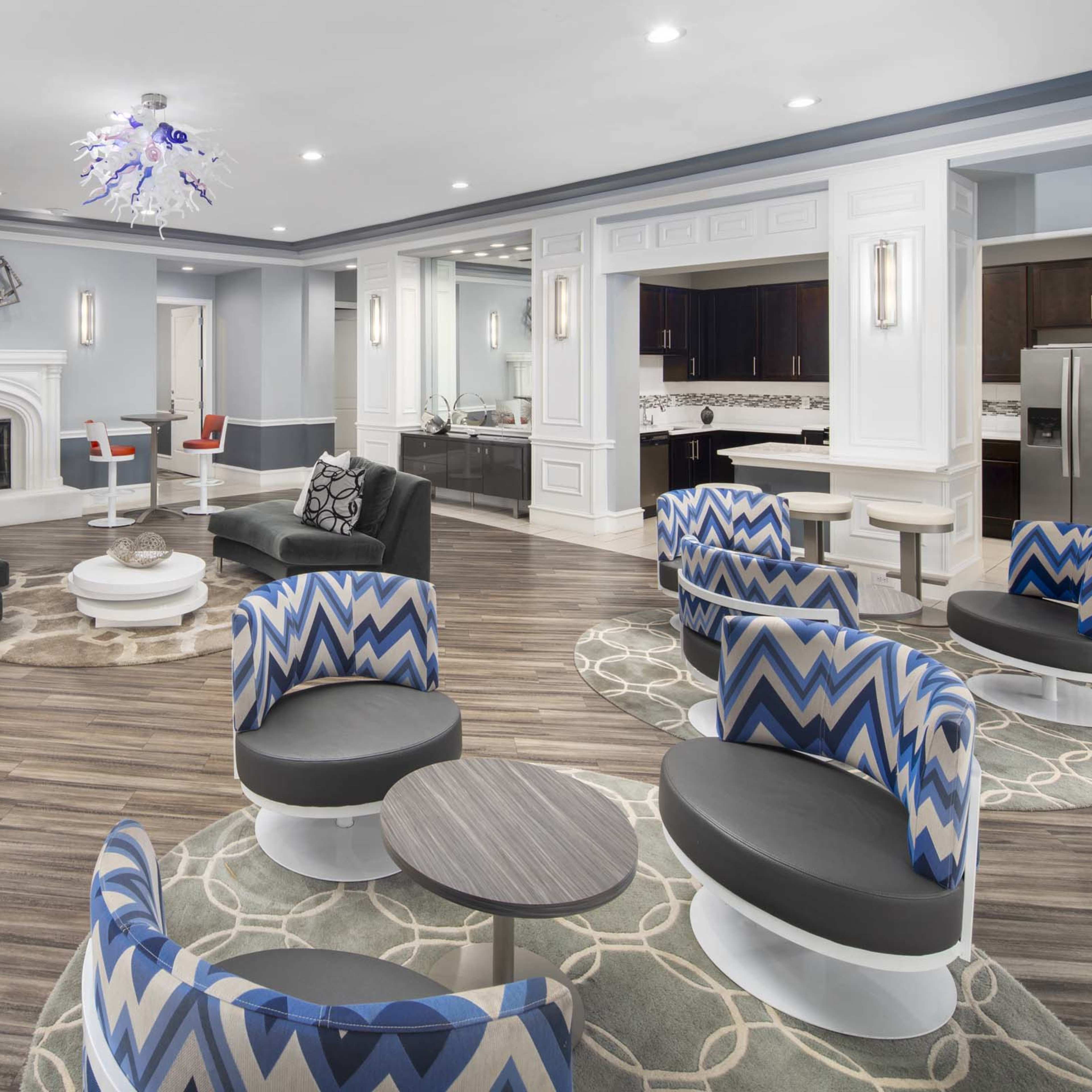 A modern lounge area featuring various seating arrangements, a fireplace, and a kitchenette in the background.
