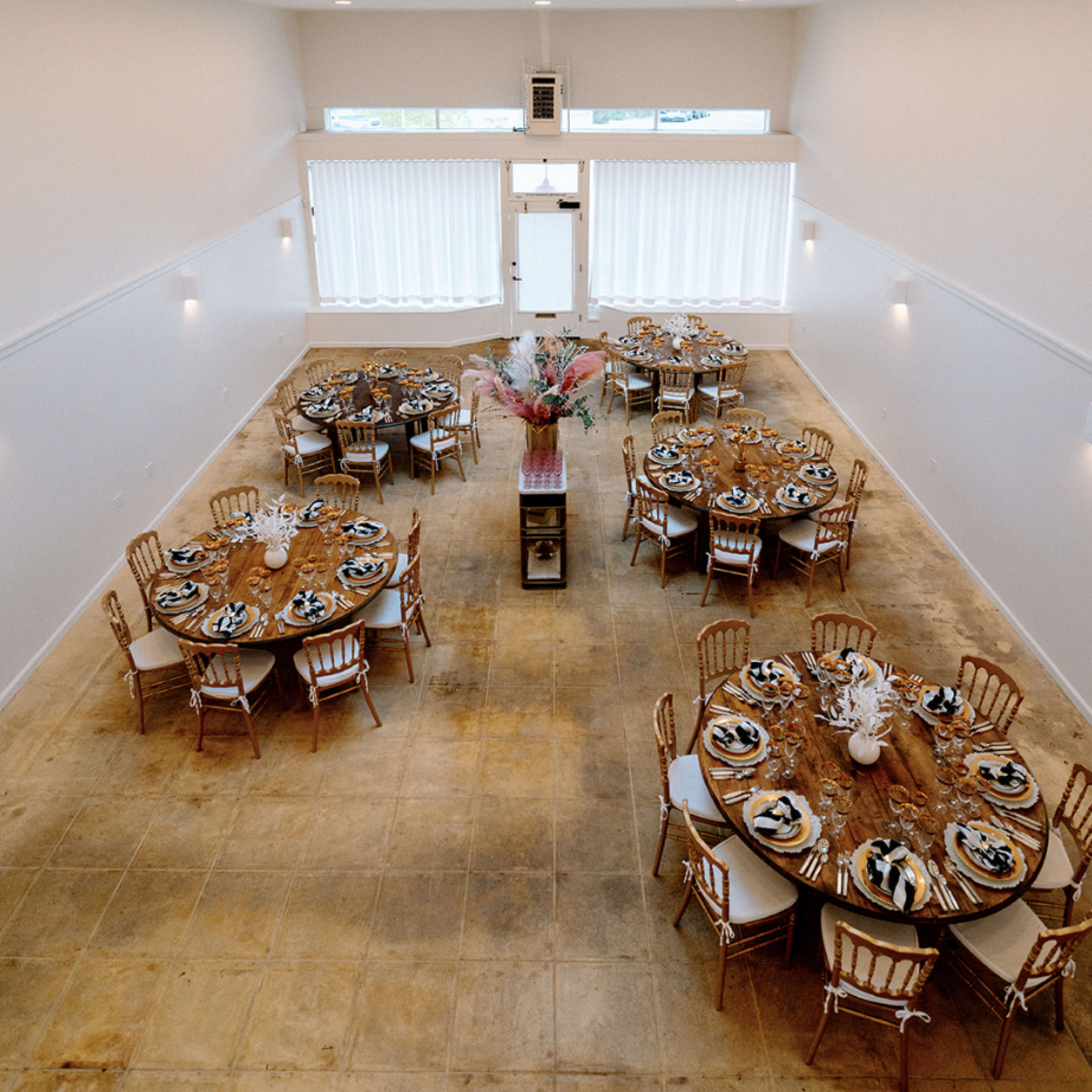 An elegantly arranged event space featuring multiple round tables set for a formal gathering, with large windows allowing natural light to enter.
