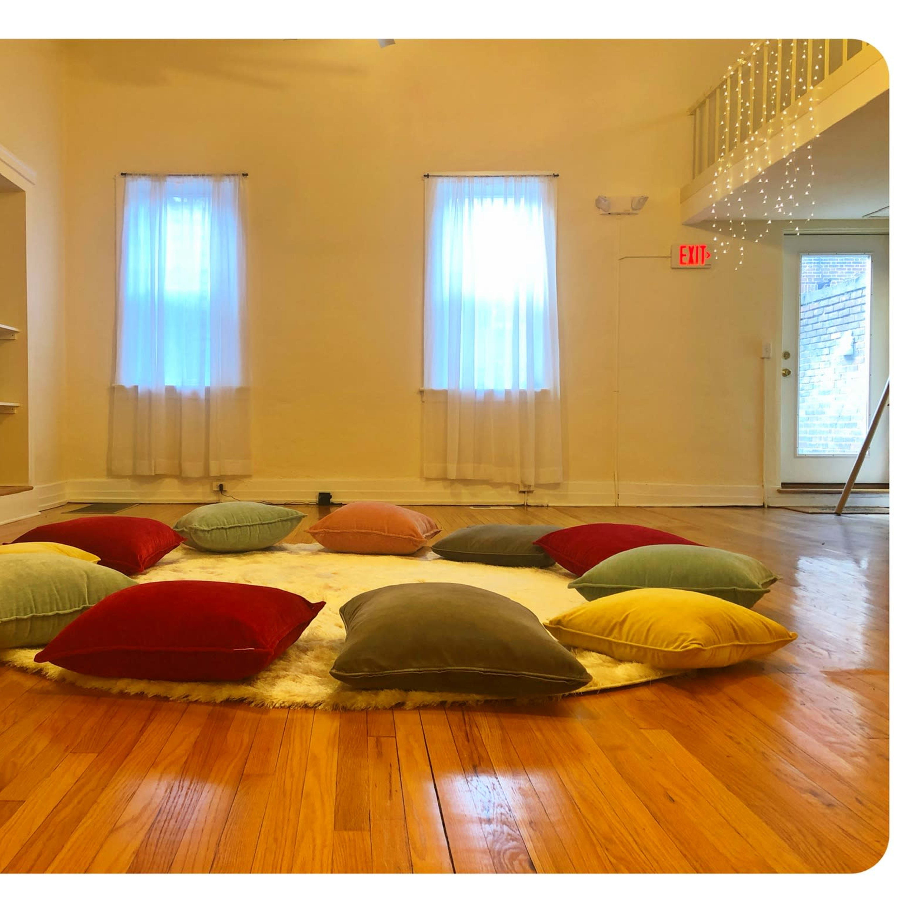 A circular arrangement of colorful pillows on a circular rug in a bright, spacious room with two windows and wooden flooring.