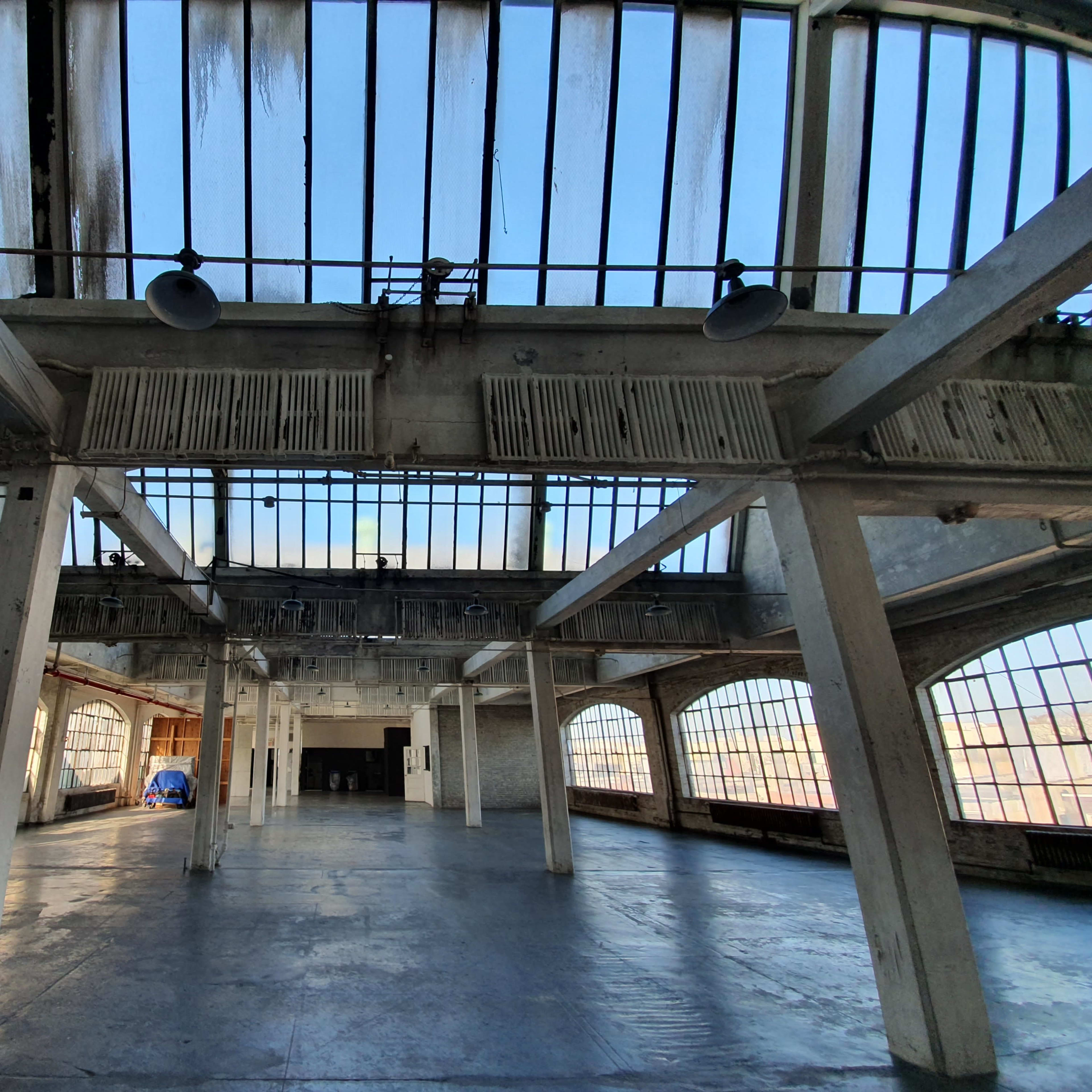 An empty industrial space with large windows and exposed beams, illuminated by natural light.