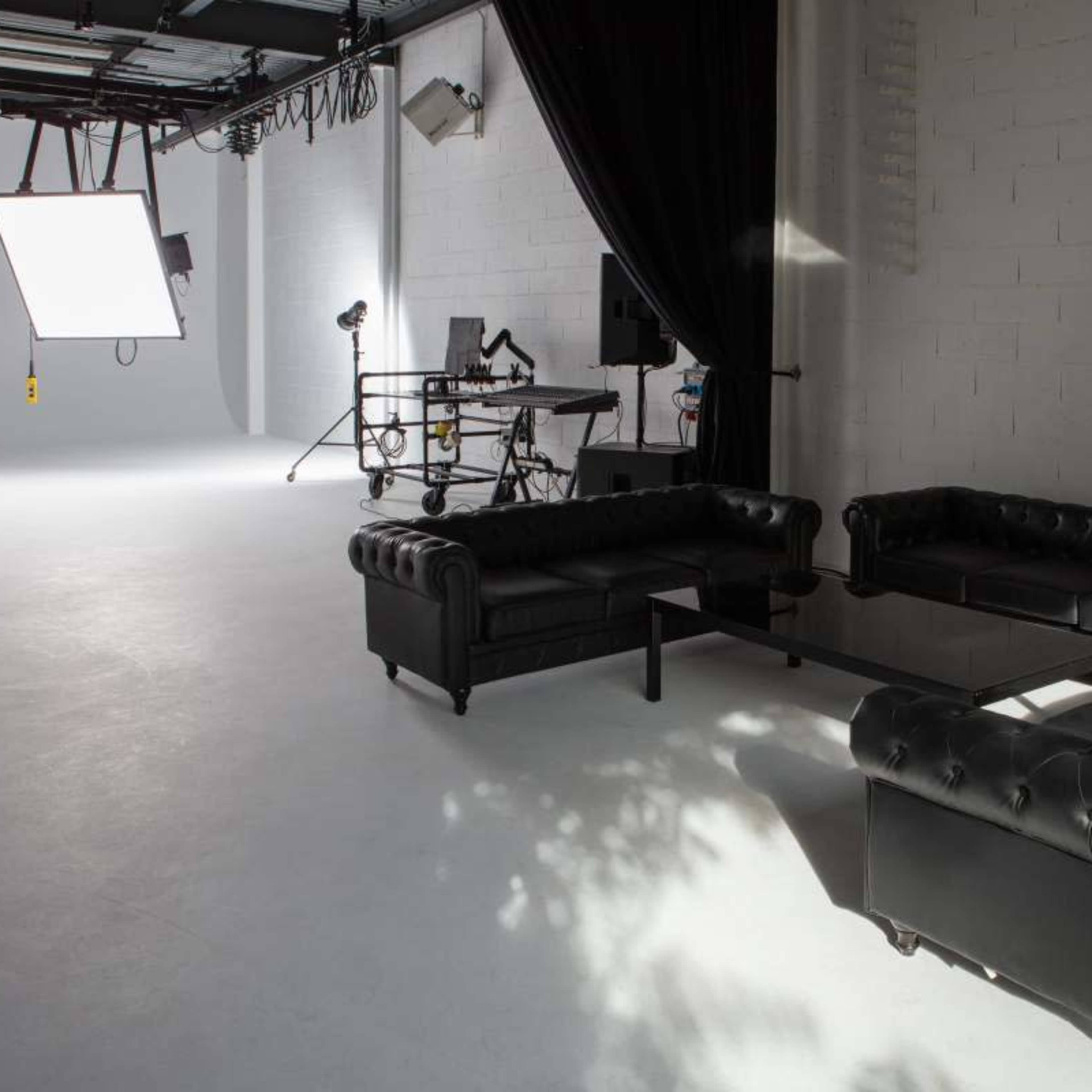 A modern photo studio features black leather couches surrounding a glass table, with professional lighting equipment and backdrops in the background.