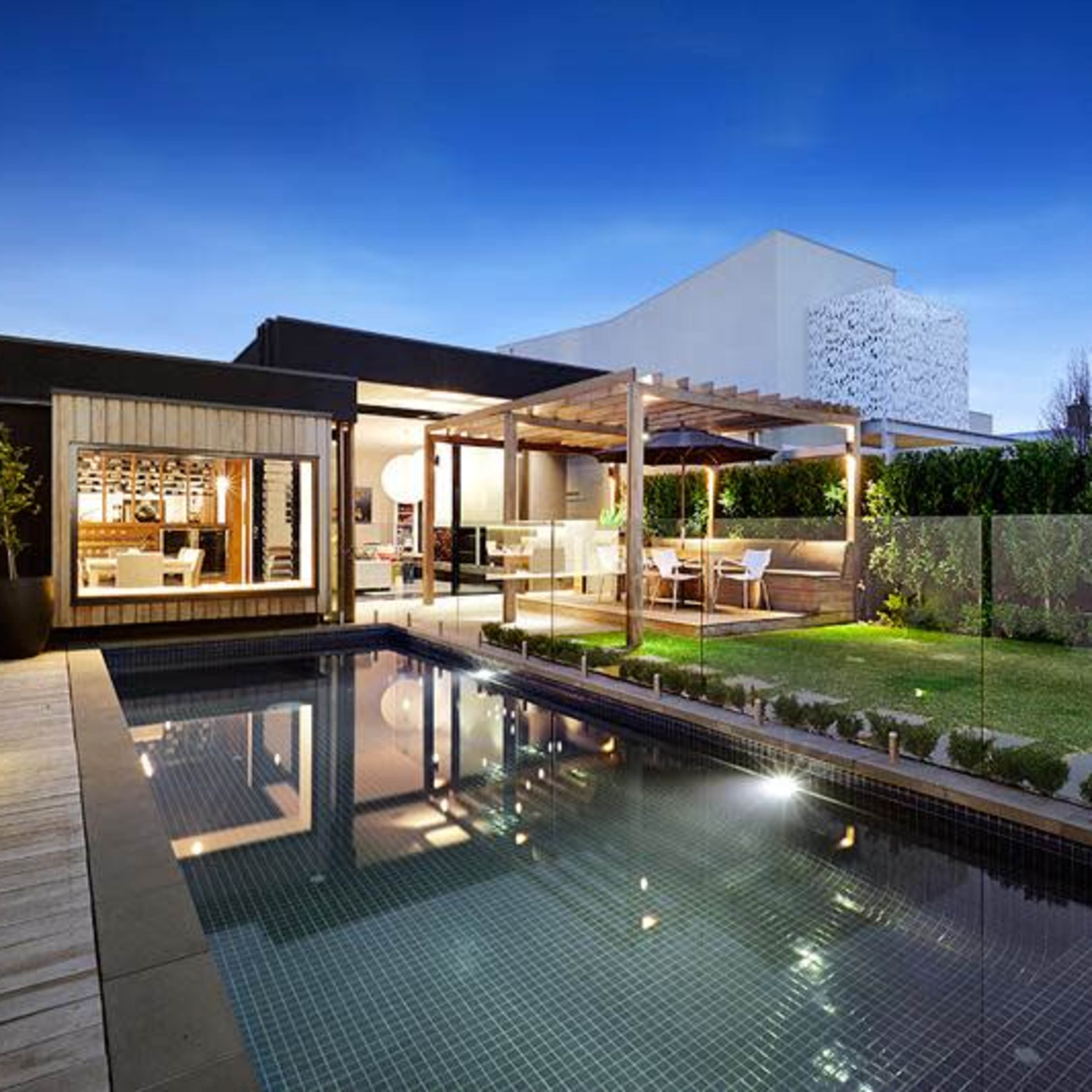 A modern backyard with a swimming pool, wooden deck, and a dining area under a pergola, surrounded by well-maintained greenery at twilight.