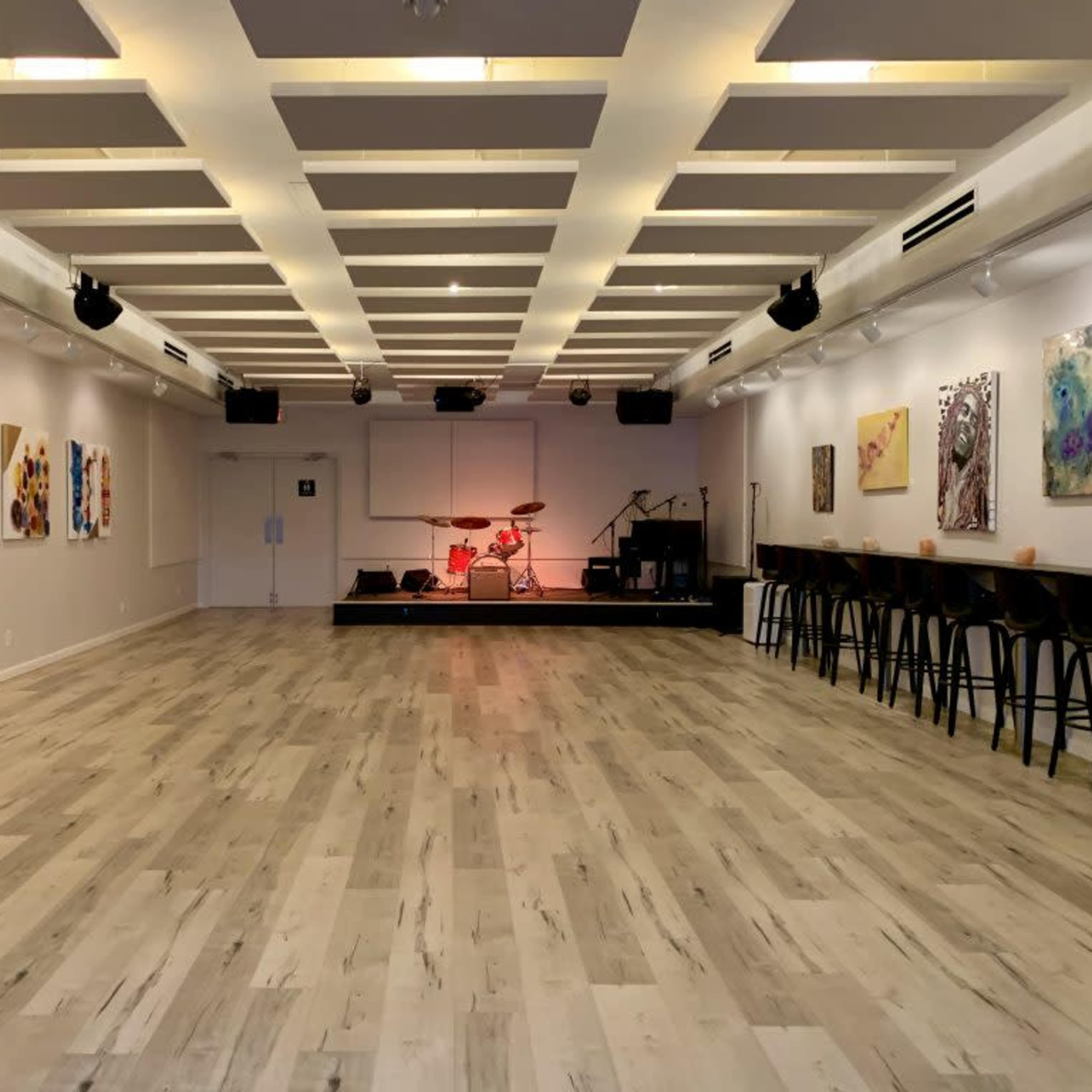 A spacious, well-lit performance area featuring a stage with a drum set, surrounded by artwork on the walls and a row of tall chairs along one side.