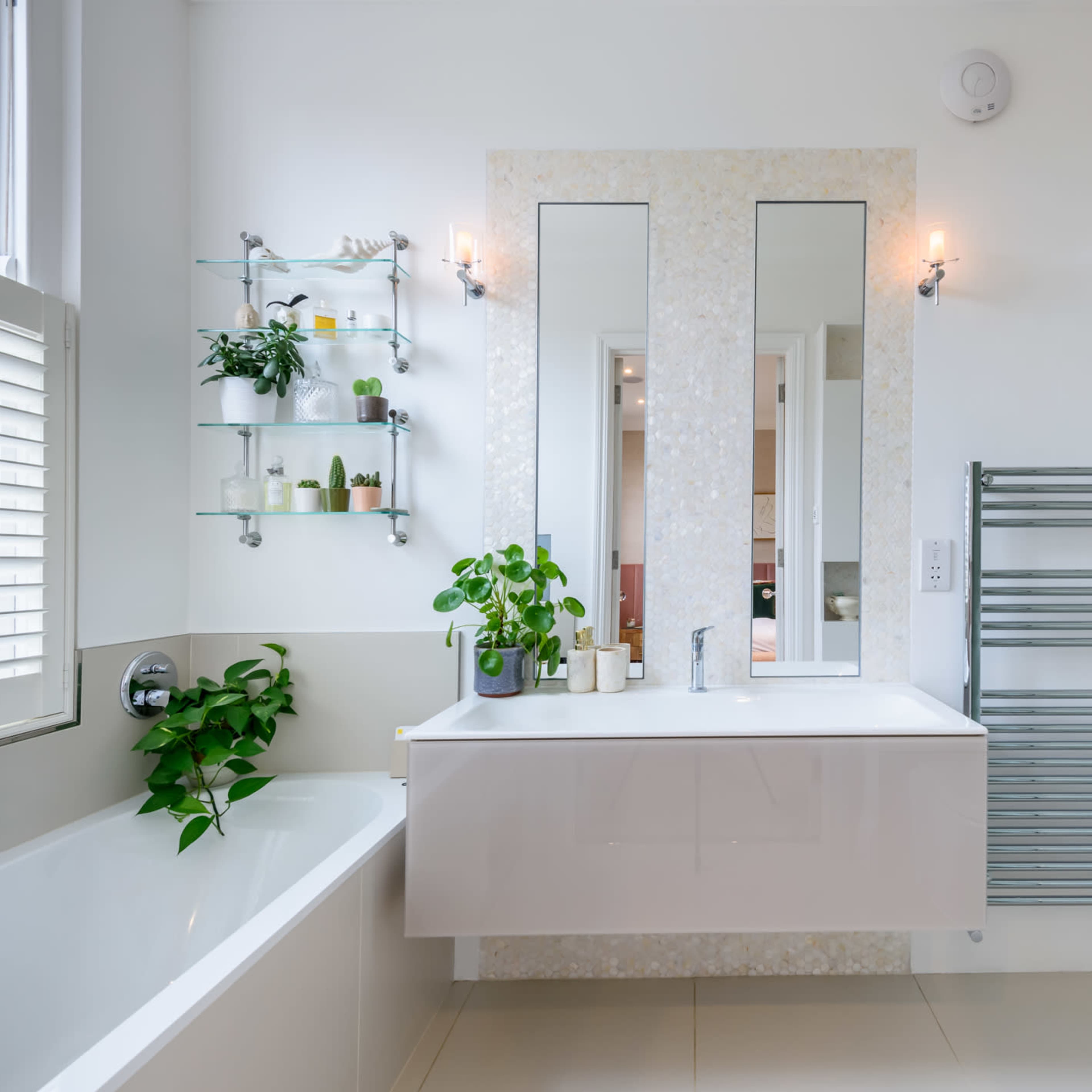 A modern bathroom featuring a bathtub, a wall-mounted vanity with two mirrors, plants on shelves, and a glass-enclosed shower.