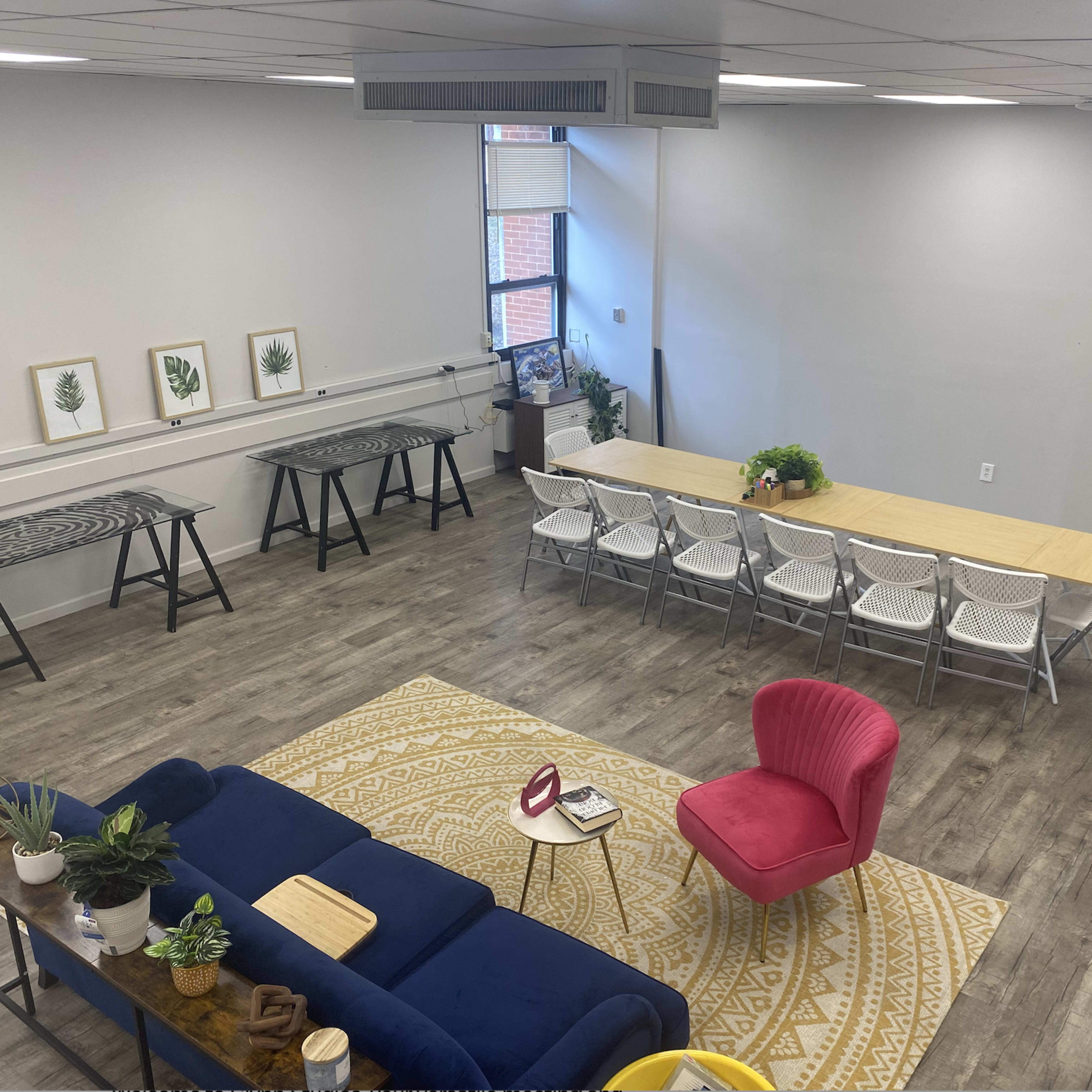 A spacious, modern communal area featuring a long dining table surrounded by chairs, a seating area with a blue sofa and a pink armchair, and several worktables against the walls.