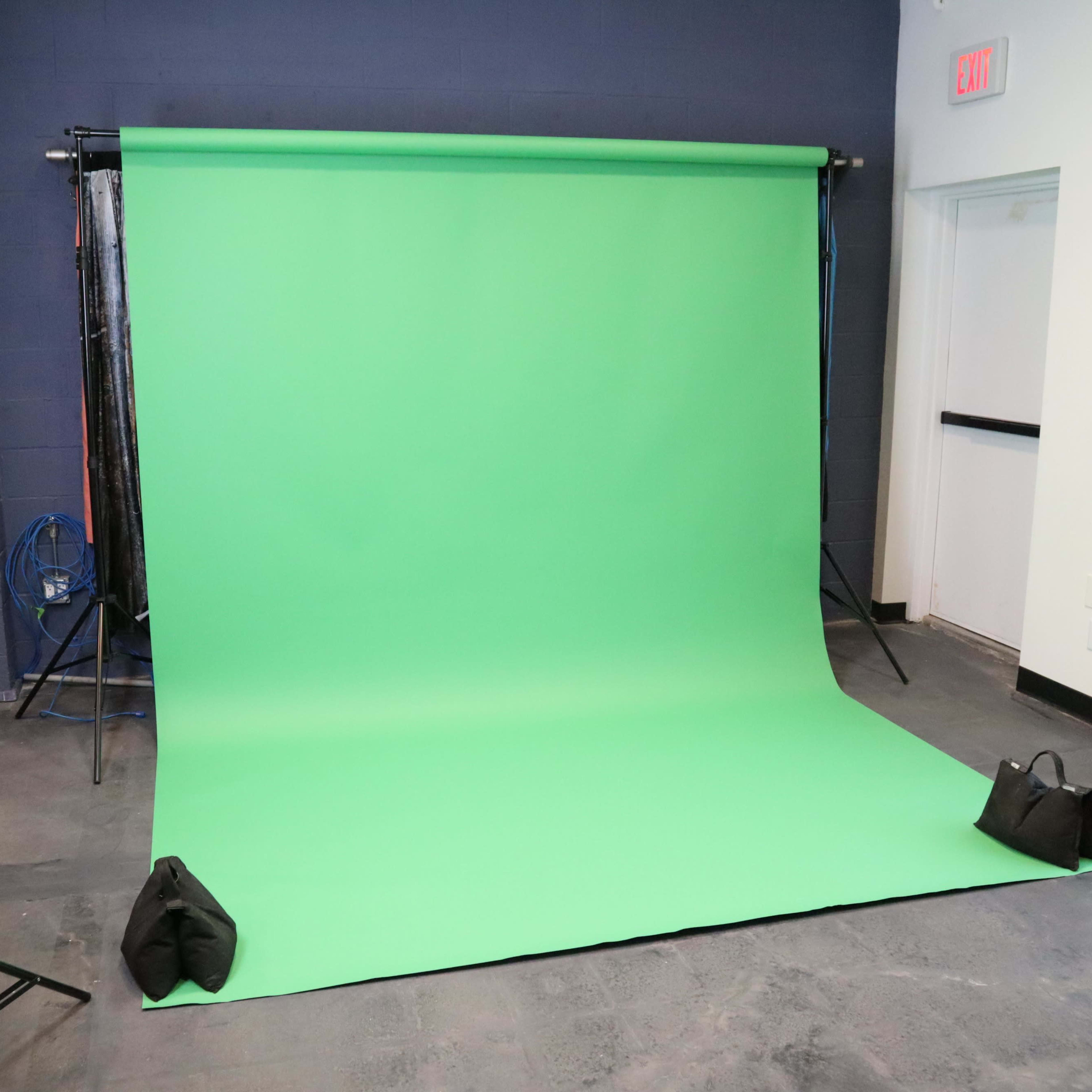 A green backdrop set up in a studio space, supported by stands and weighted down by bags at the base.