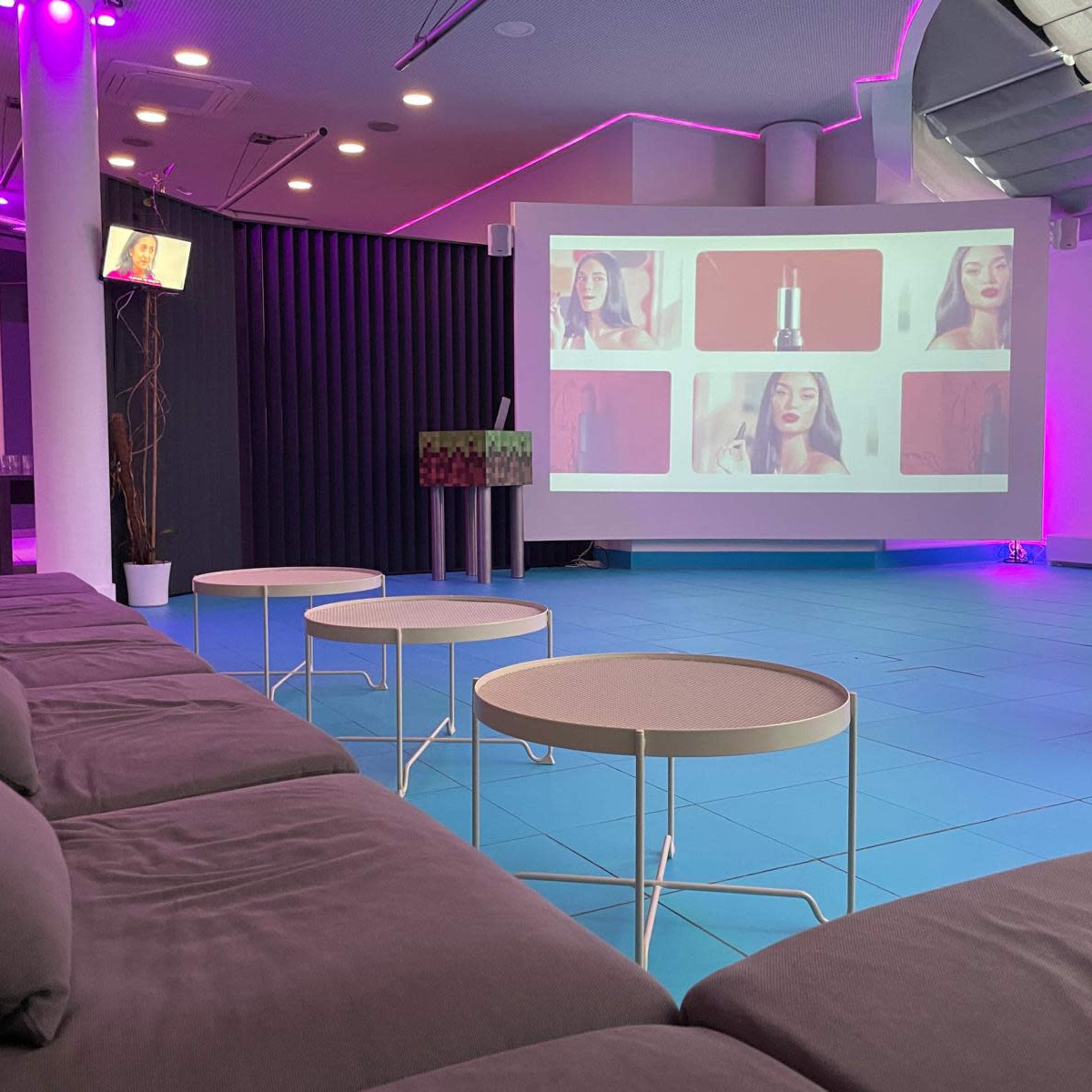 A modern lounge area featuring gray couches and white tables, with a large screen projecting a video and ambient purple lighting.