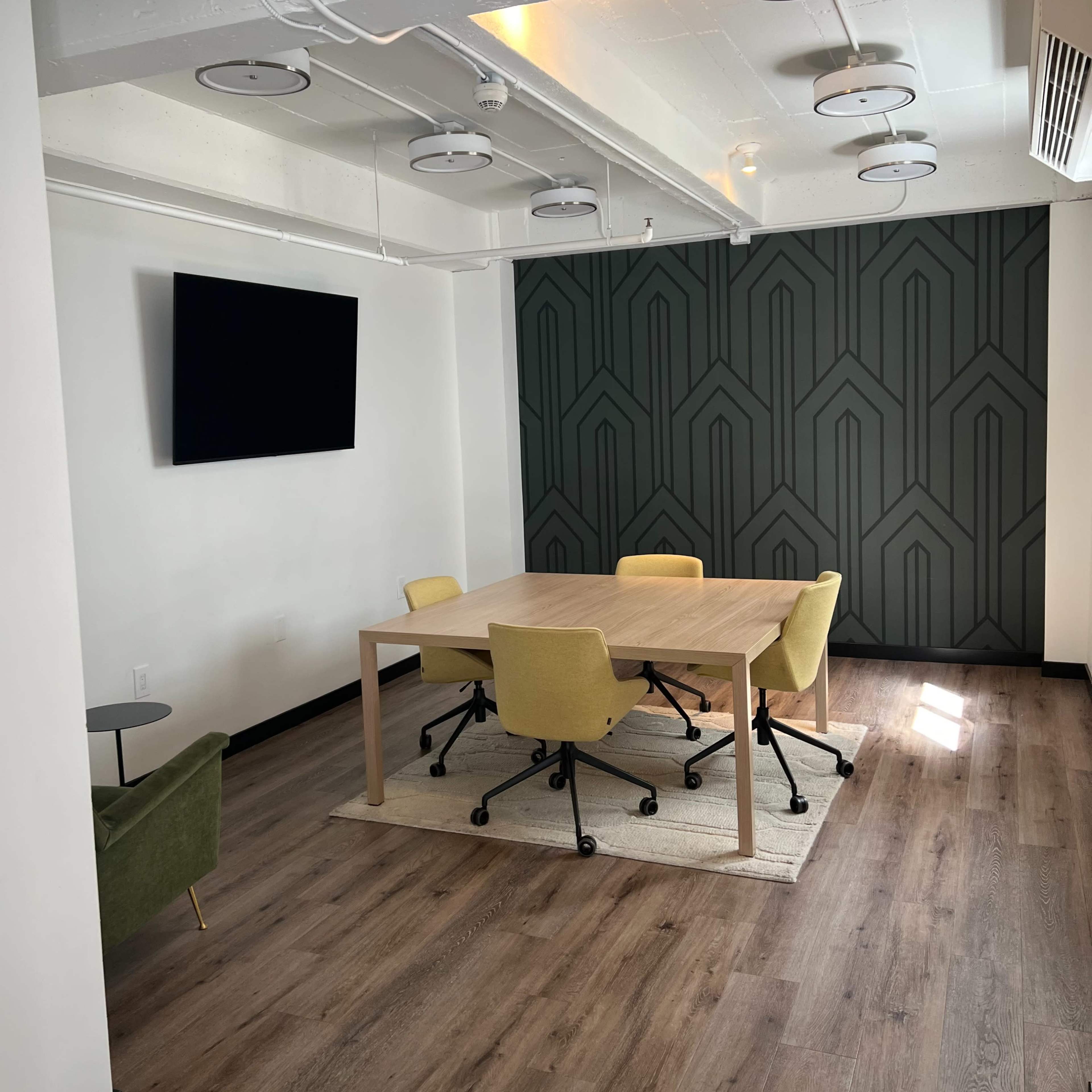 A modern meeting room features a wooden table surrounded by yellow chairs, with a large TV mounted on a wall and a textured green accent wall in the background.
