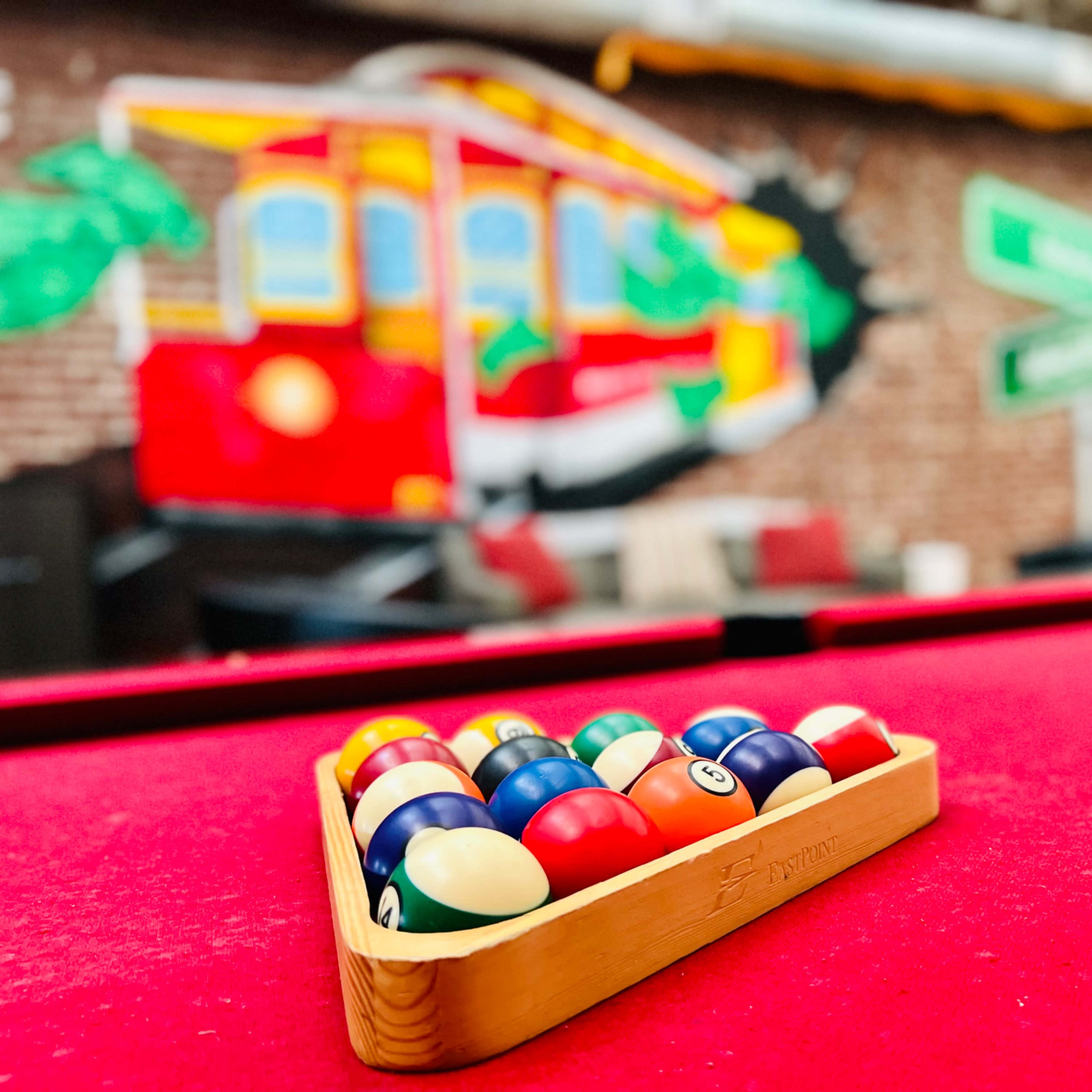 A triangle of billiard balls rests on a red felt pool table in front of a vibrant mural featuring a red streetcar.