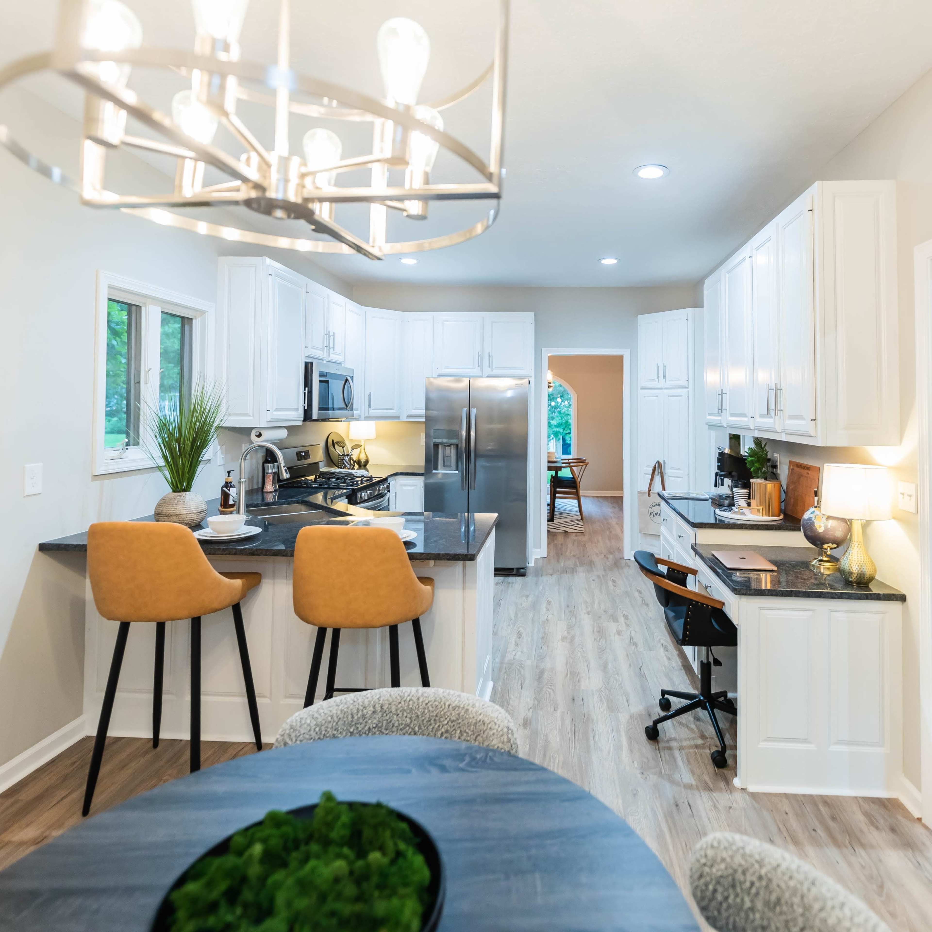 A modern kitchen and dining area featuring white cabinets, a central island with two chairs, and a visible workspace leading to another room.