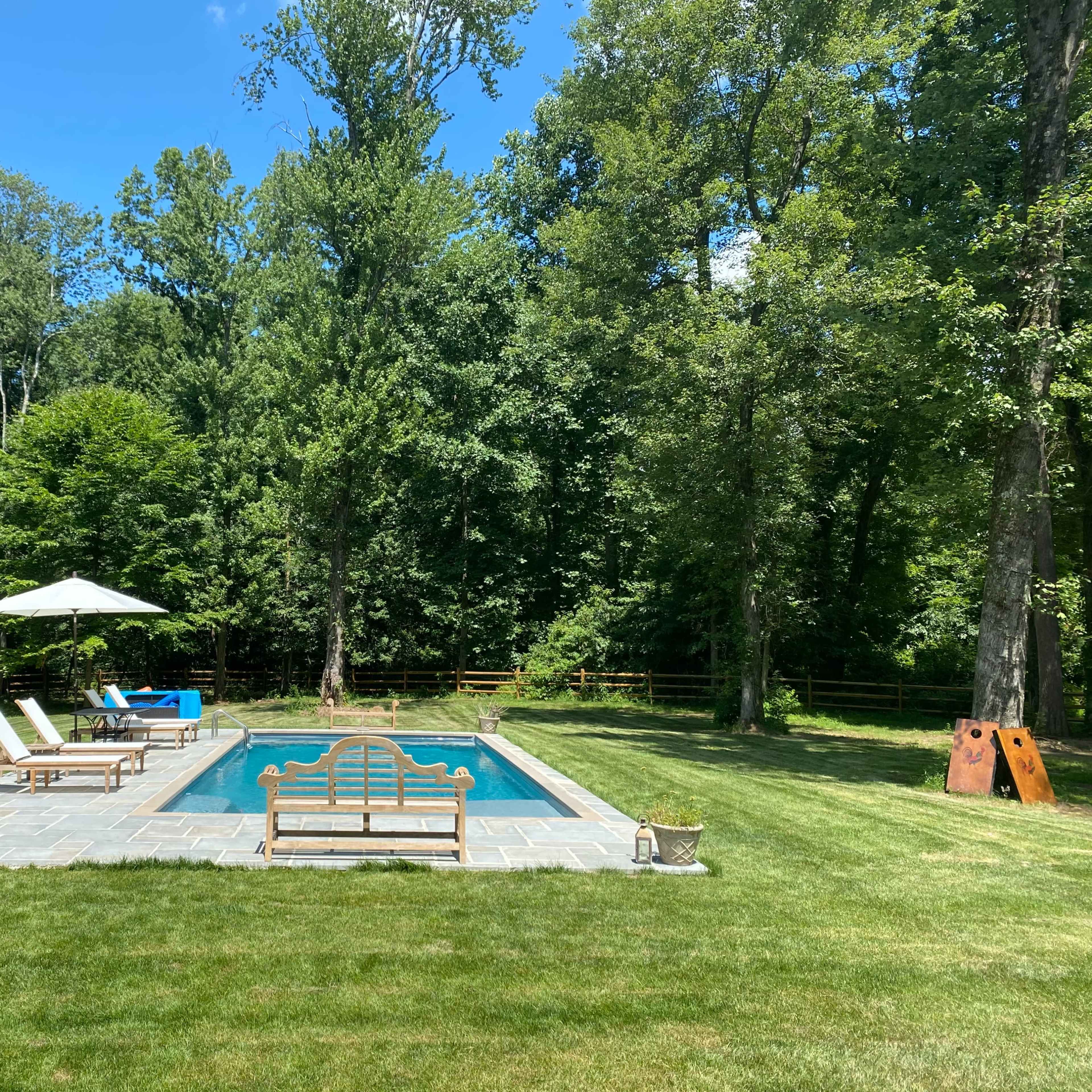 A rectangular swimming pool is surrounded by wooden lounge chairs and an umbrella, with a grassy yard and trees in the background.