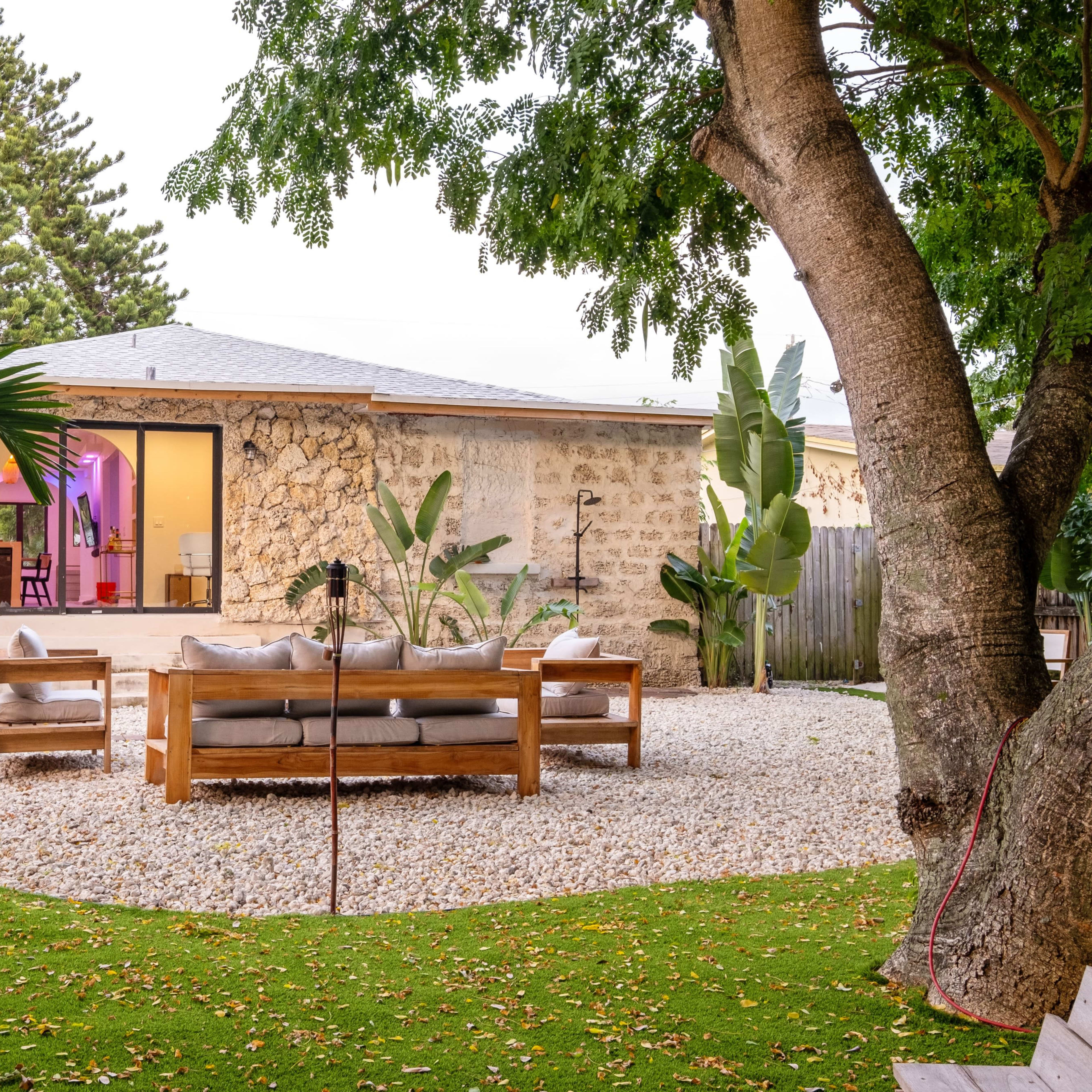 A wooden seating area is set on gravel in a landscaped yard with tropical plants, facing a stone house.