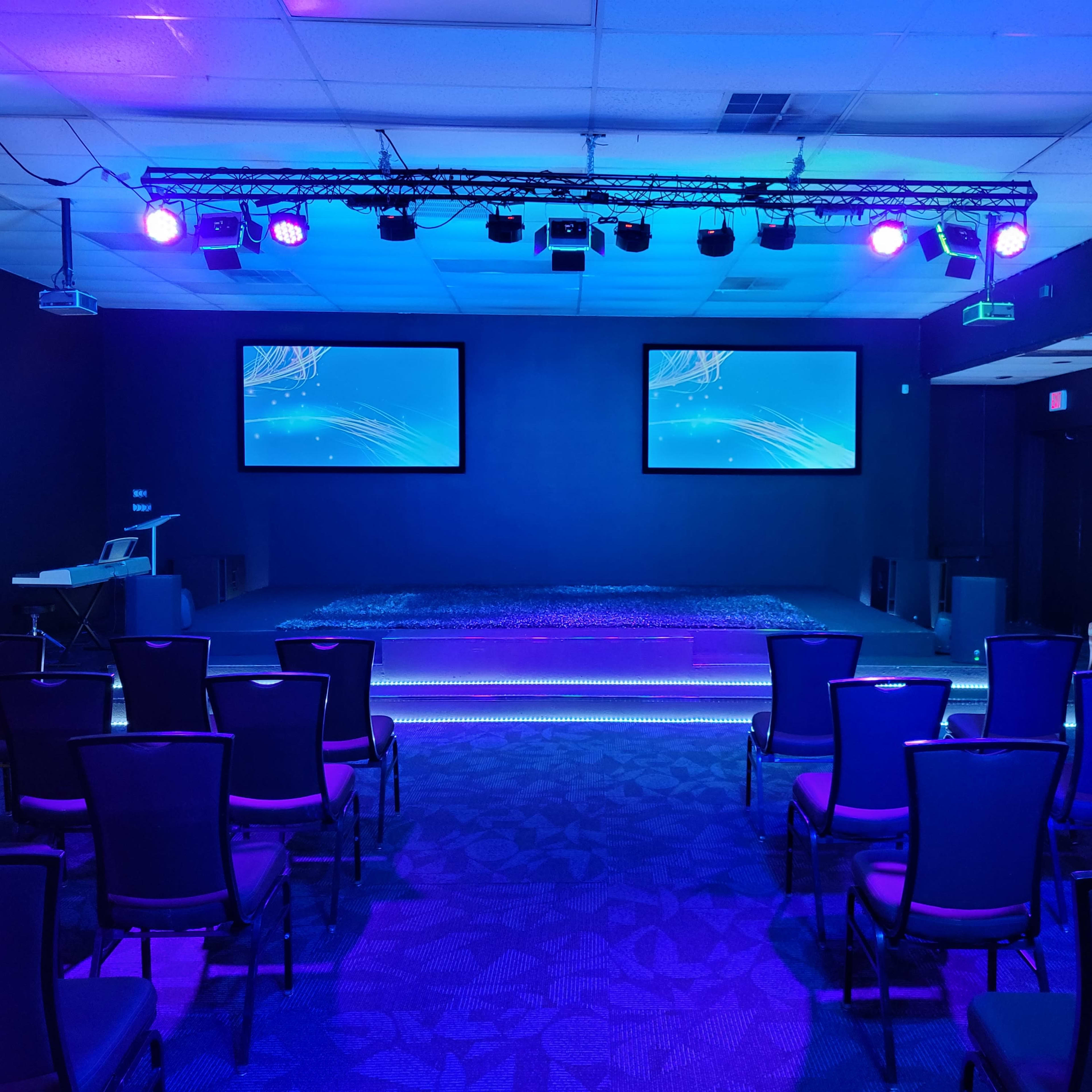A conference room with two screens, purple lighting, and arranged chairs facing a raised stage area.