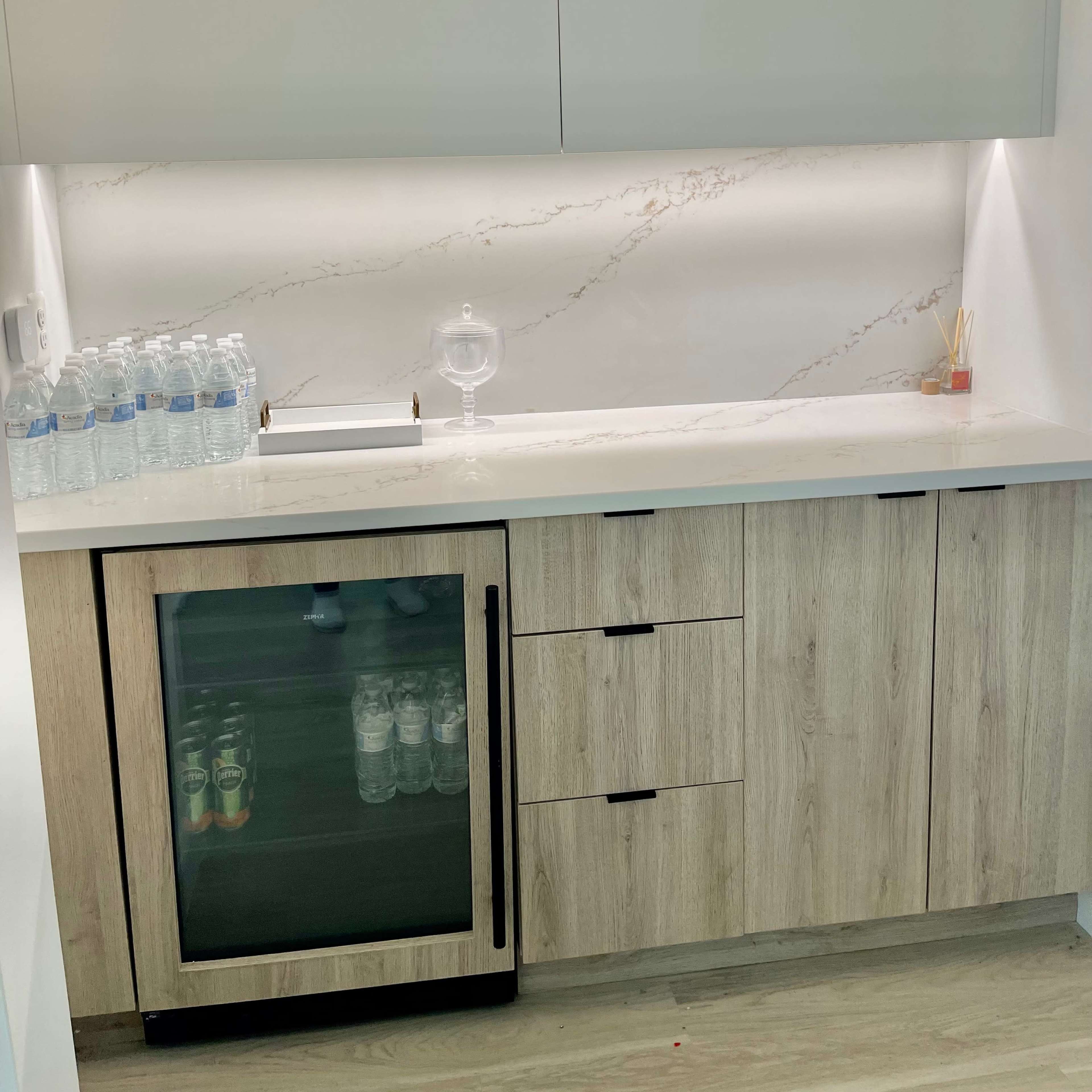 A modern kitchenette with a countertop, a glass cabinet containing drinks, and a row of bottled water on the countertop.