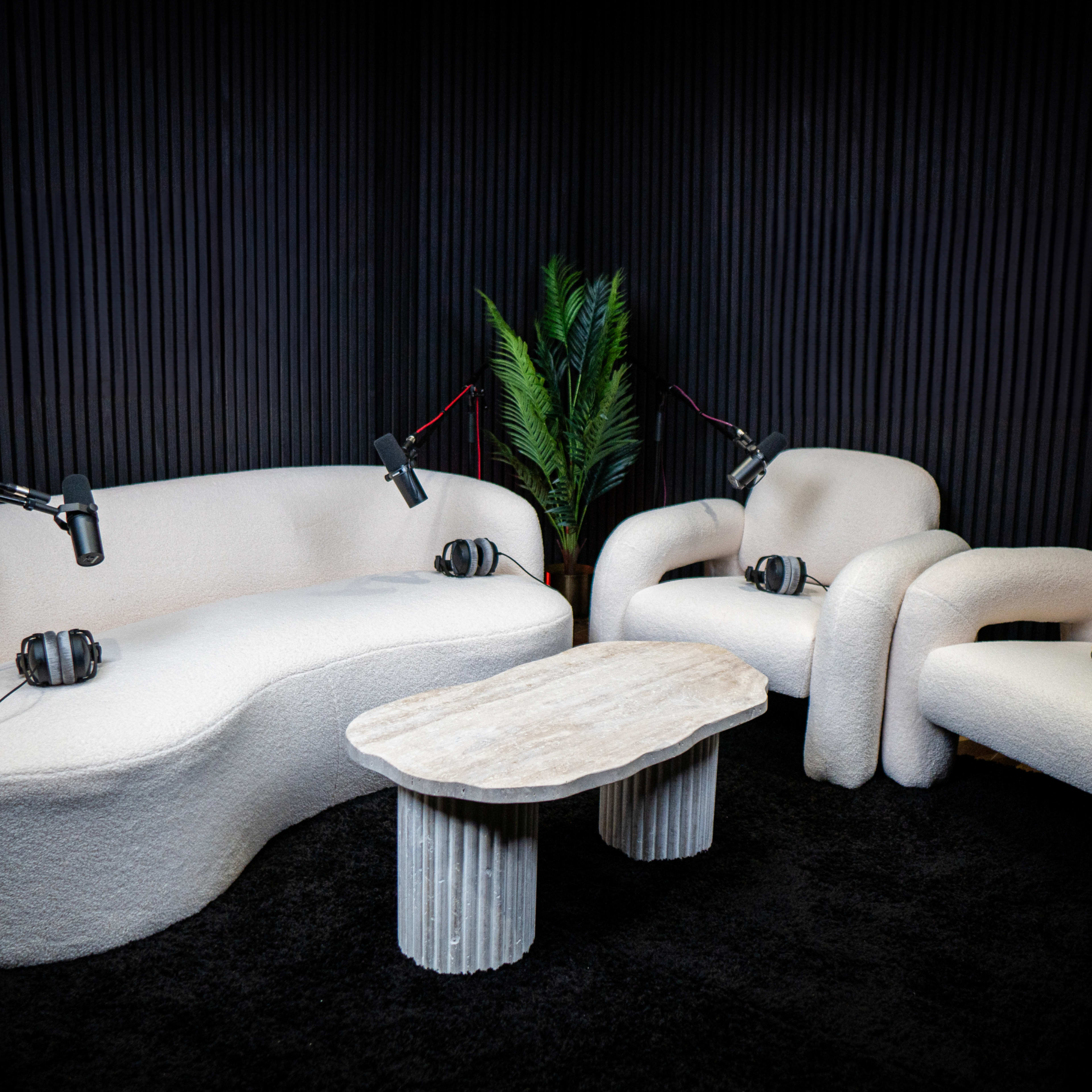A cozy audio recording setup featuring a white, curved sofa and two matching armchairs, all with microphones and headphones positioned on them, alongside a stone coffee table.