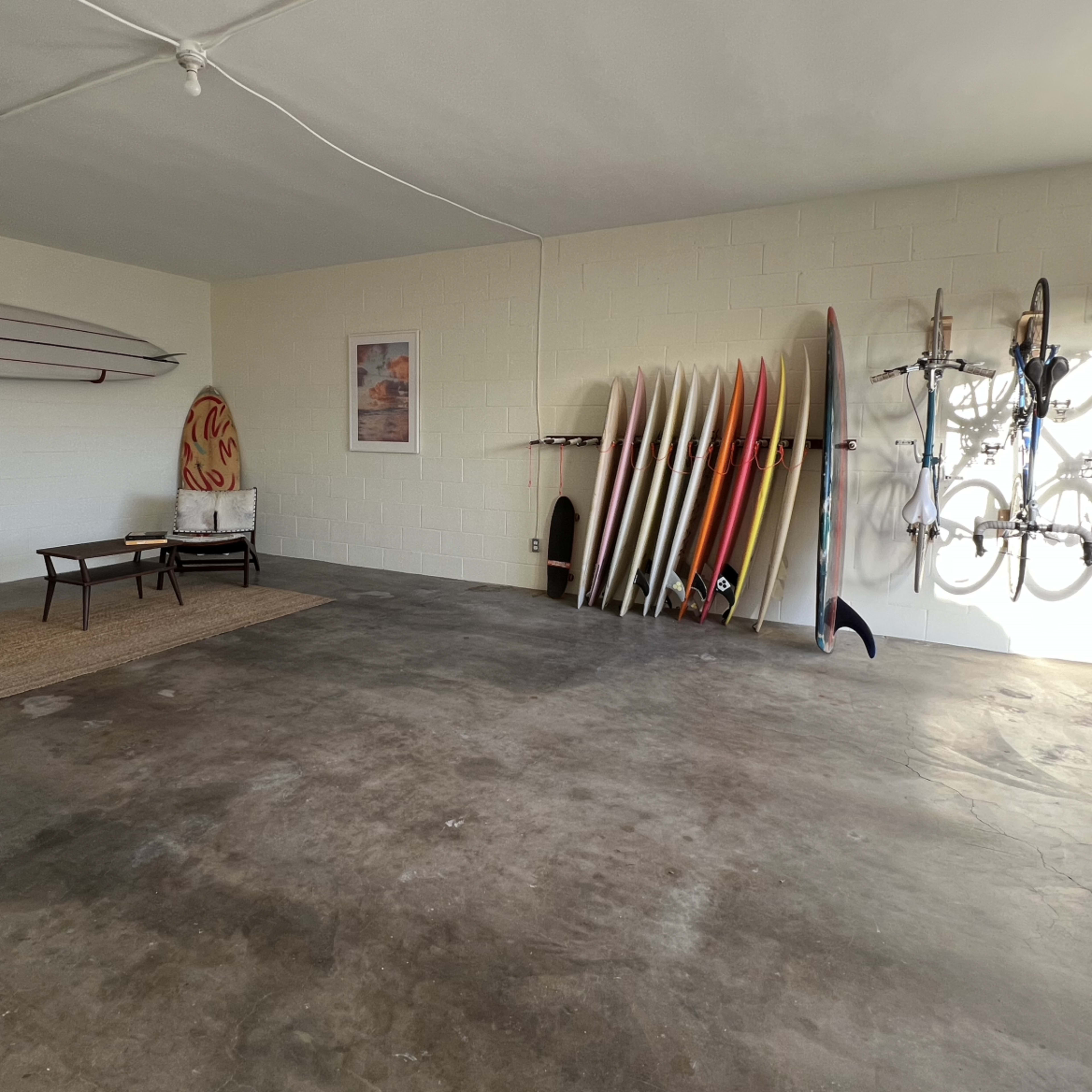 A spacious garage with surfboards and bicycles neatly arranged on the walls and a seating area consisting of a chair and small table.