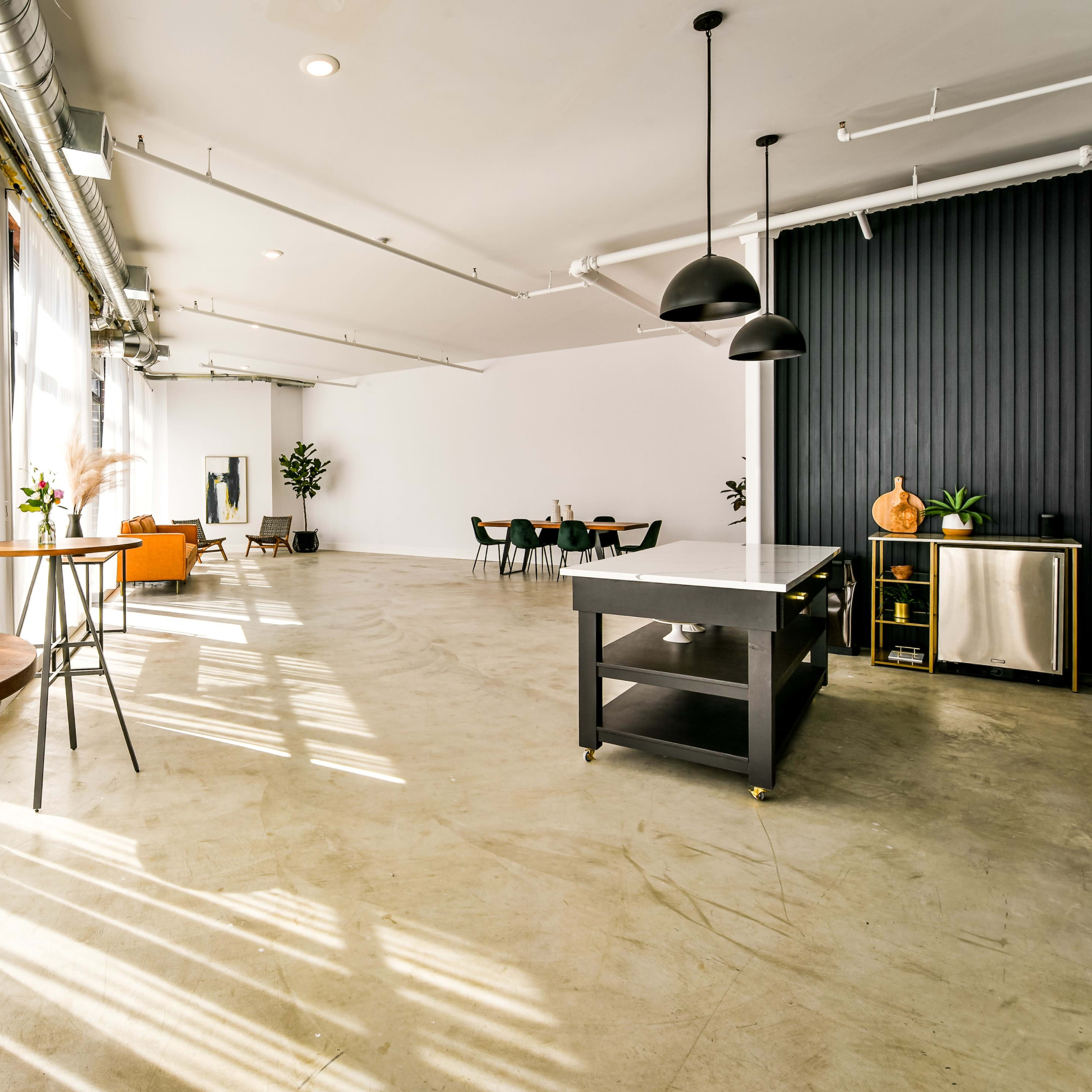 A spacious, minimalist interior with large windows, natural light flooding in, and a mix of wooden tables, green chairs, and a dark feature wall.