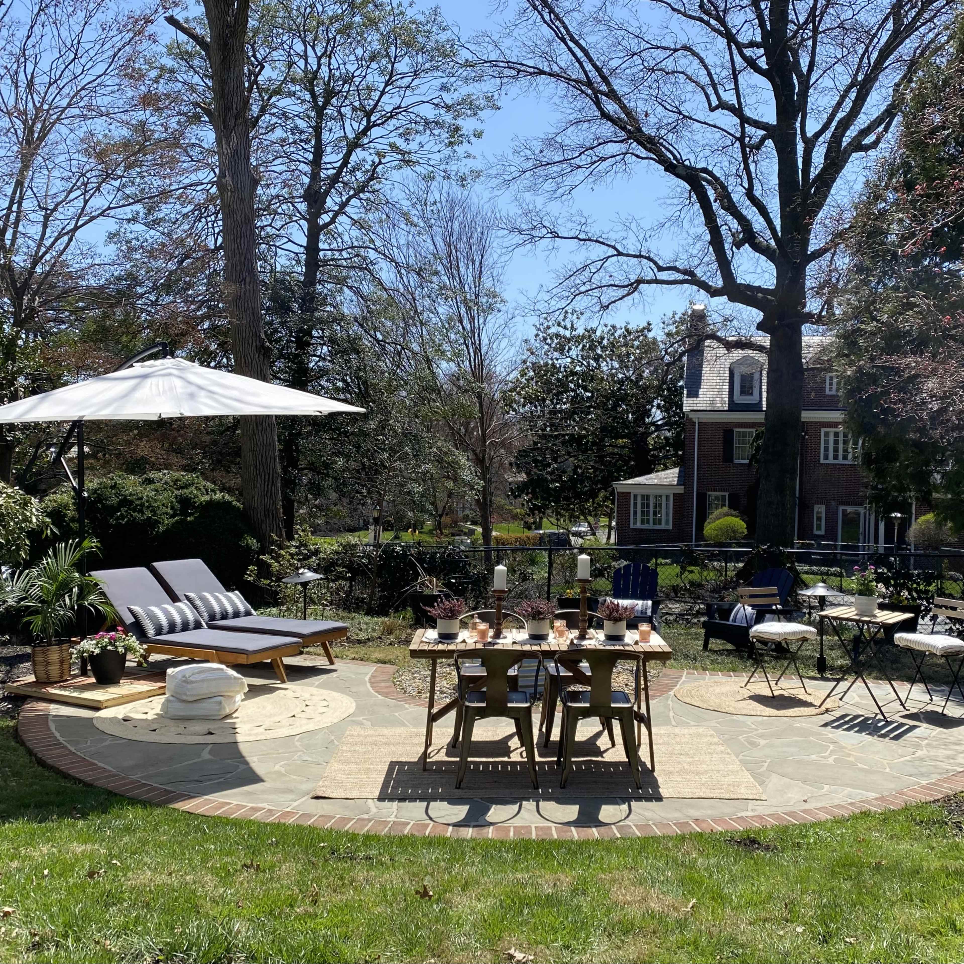 A spacious outdoor patio features a dining table set for several guests, lounge chairs, an umbrella, and greenery surrounding a stone circular area.