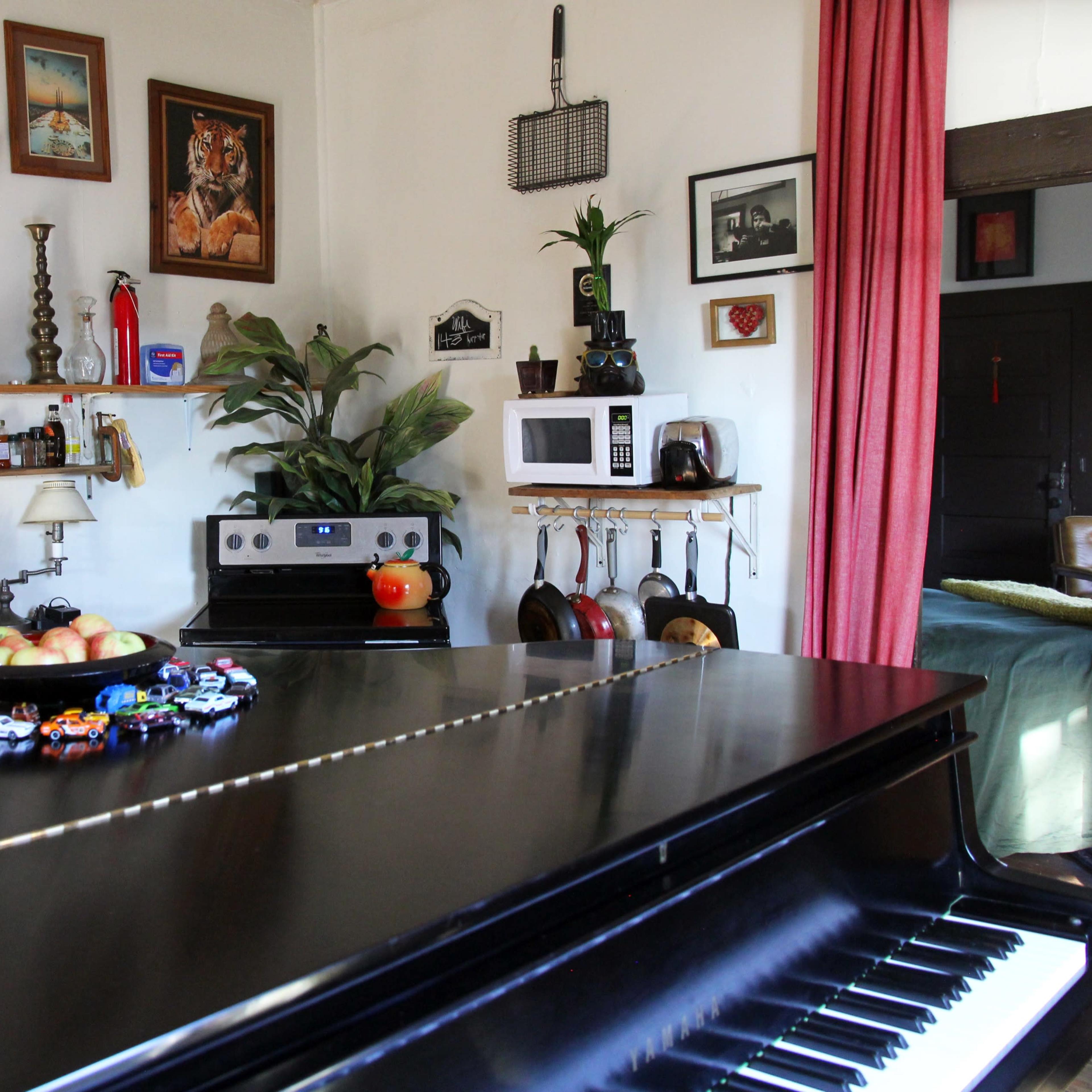 A black grand piano is positioned in the foreground of a cozy kitchen and living area, decorated with various plants, artwork, and kitchen utensils.