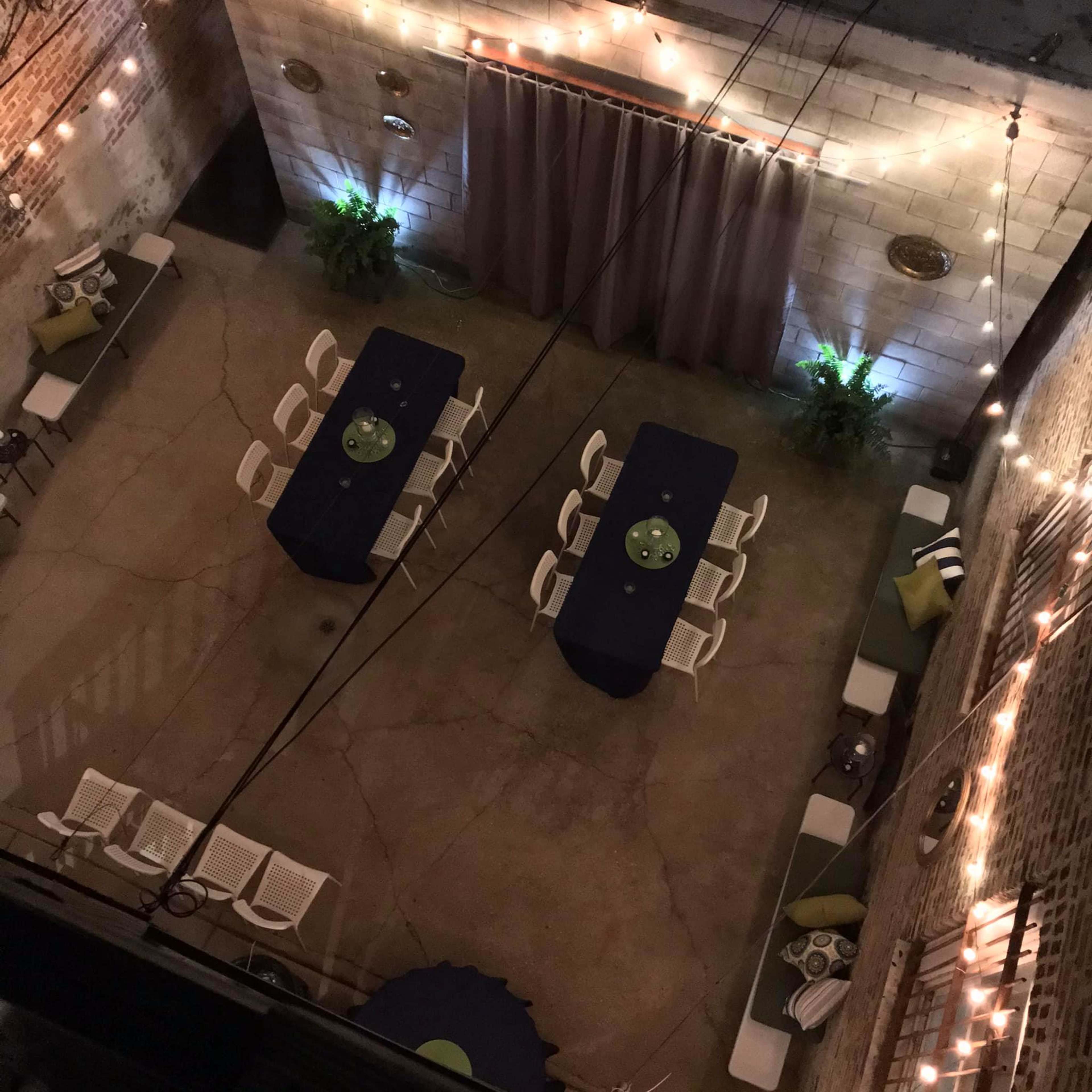 An outdoor patio area with two long tables, white chairs, and string lights illuminating the brick walls.