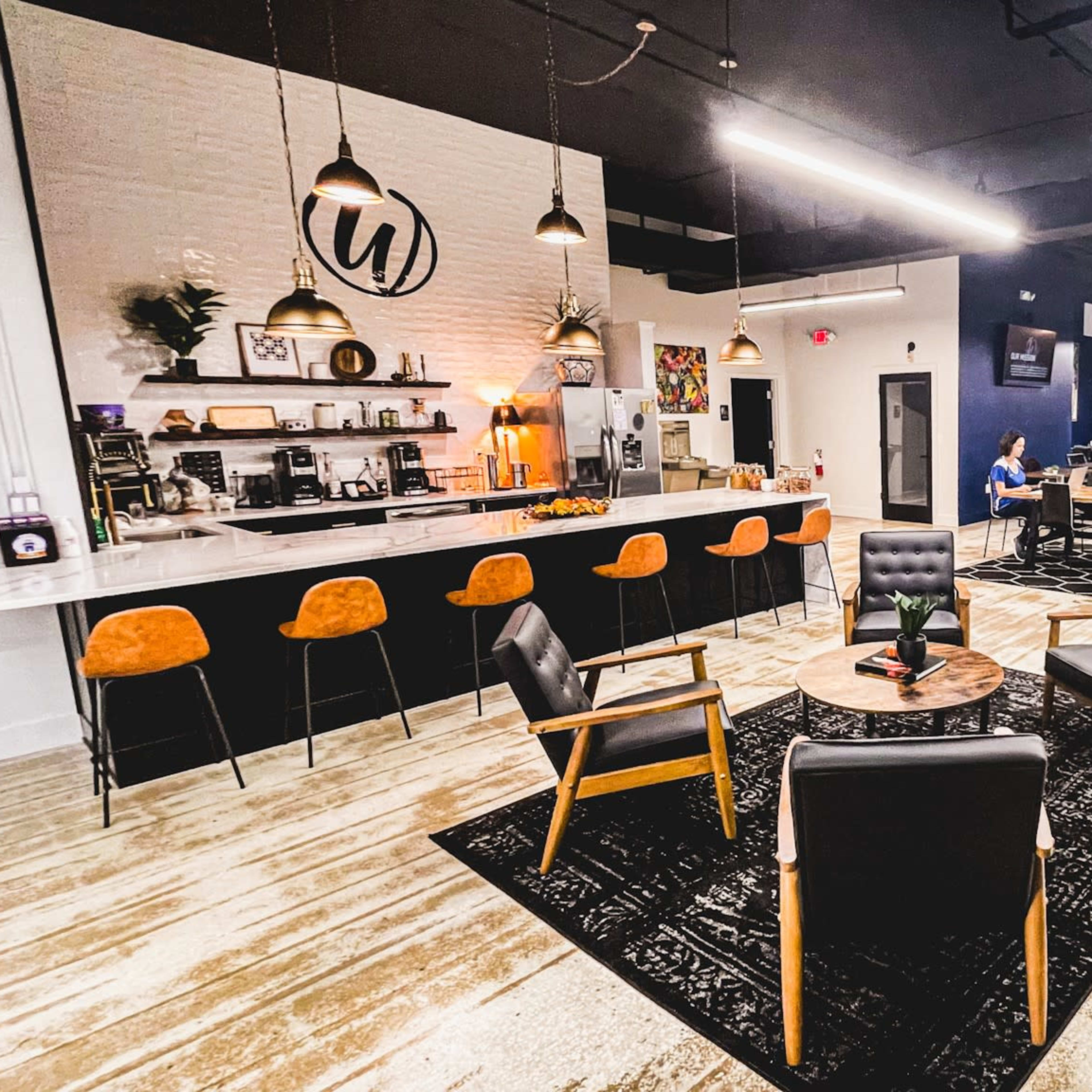 A modern co-working space with a seating area featuring black chairs and a round table, a kitchen counter with orange bar stools, and various plants in the background.