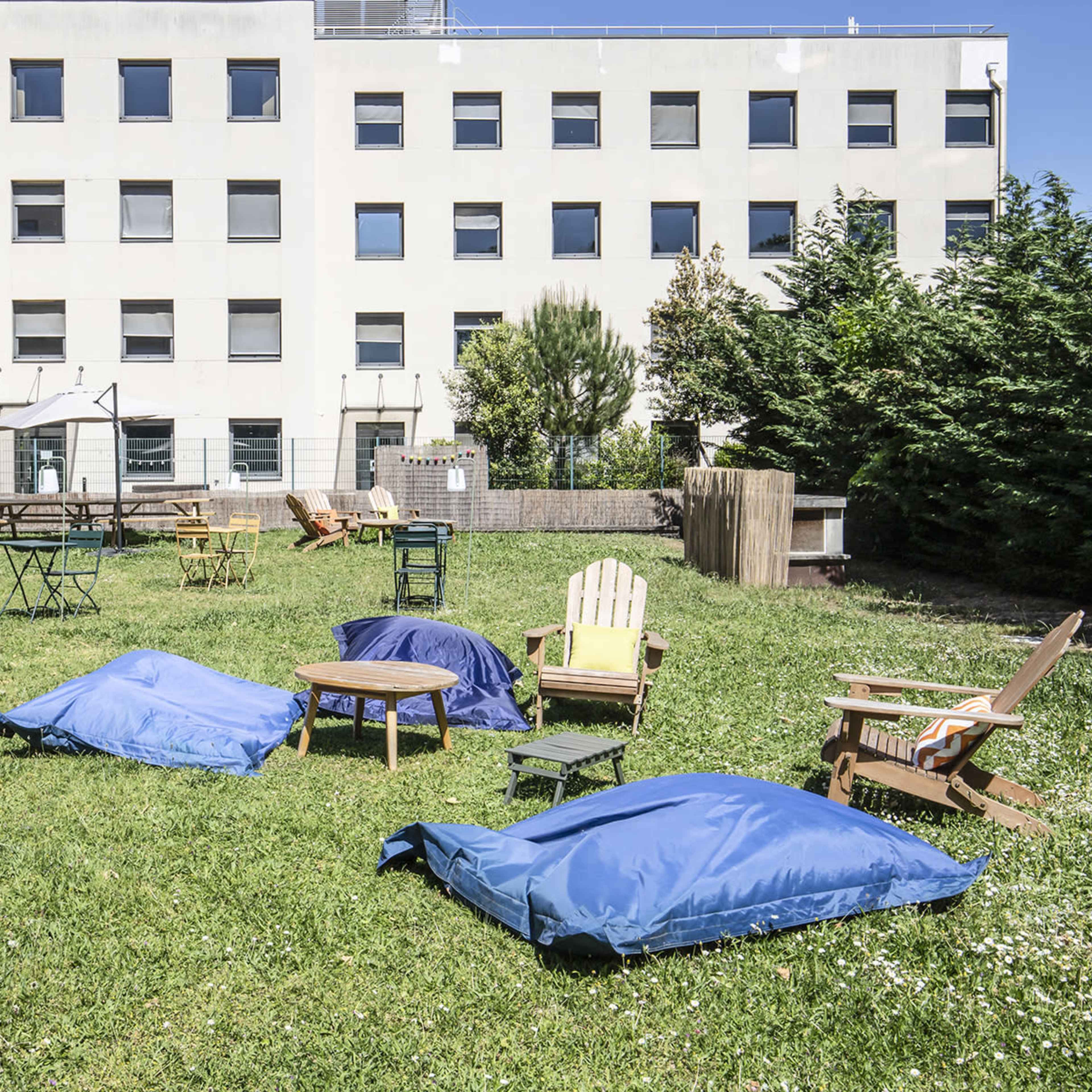 A grassy outdoor area with various seating arrangements, including blue bean bags, wooden chairs, and tables, adjacent to a white building.