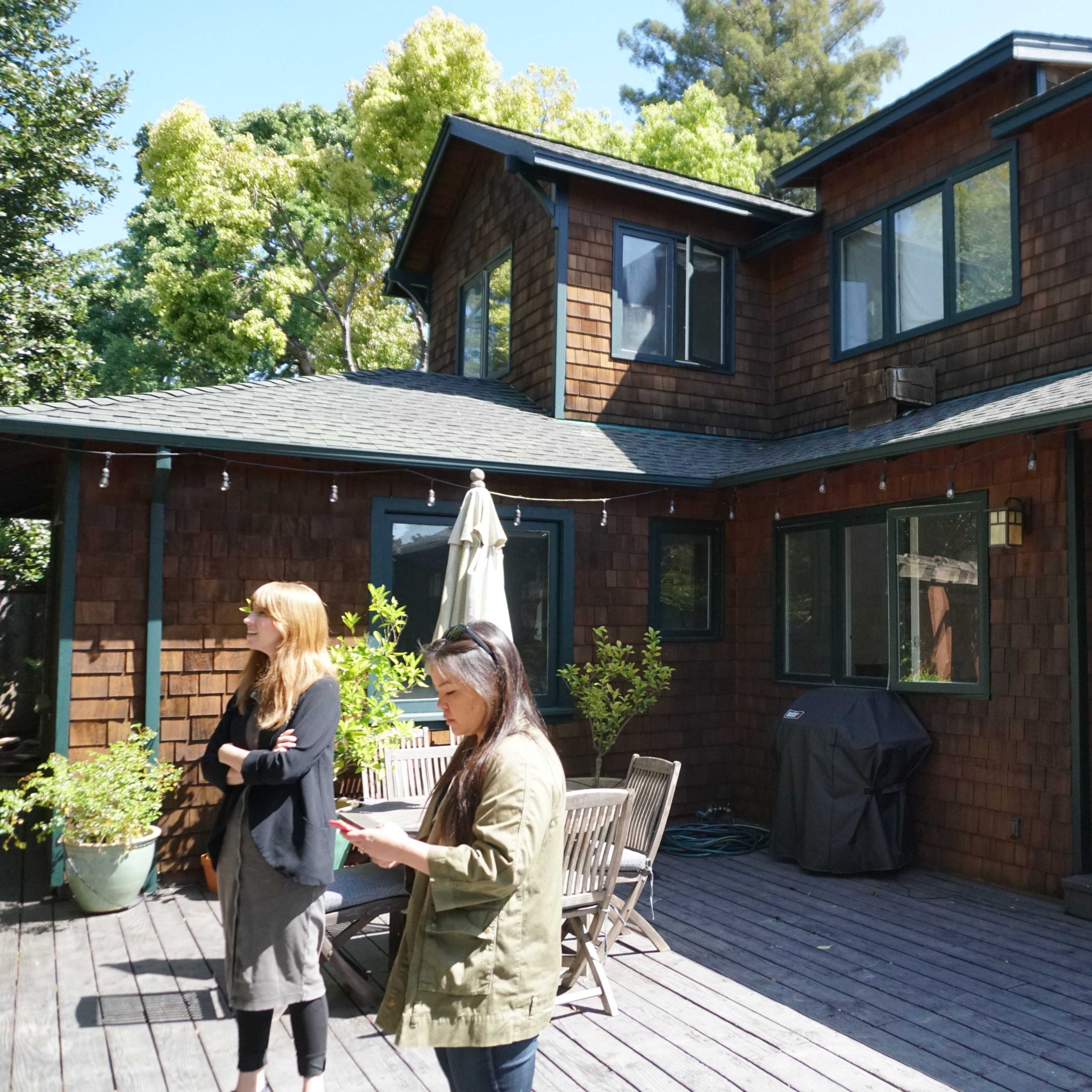 Two women stand on a wooden deck in front of a house featuring a multi-level, shingled exterior and large windows.