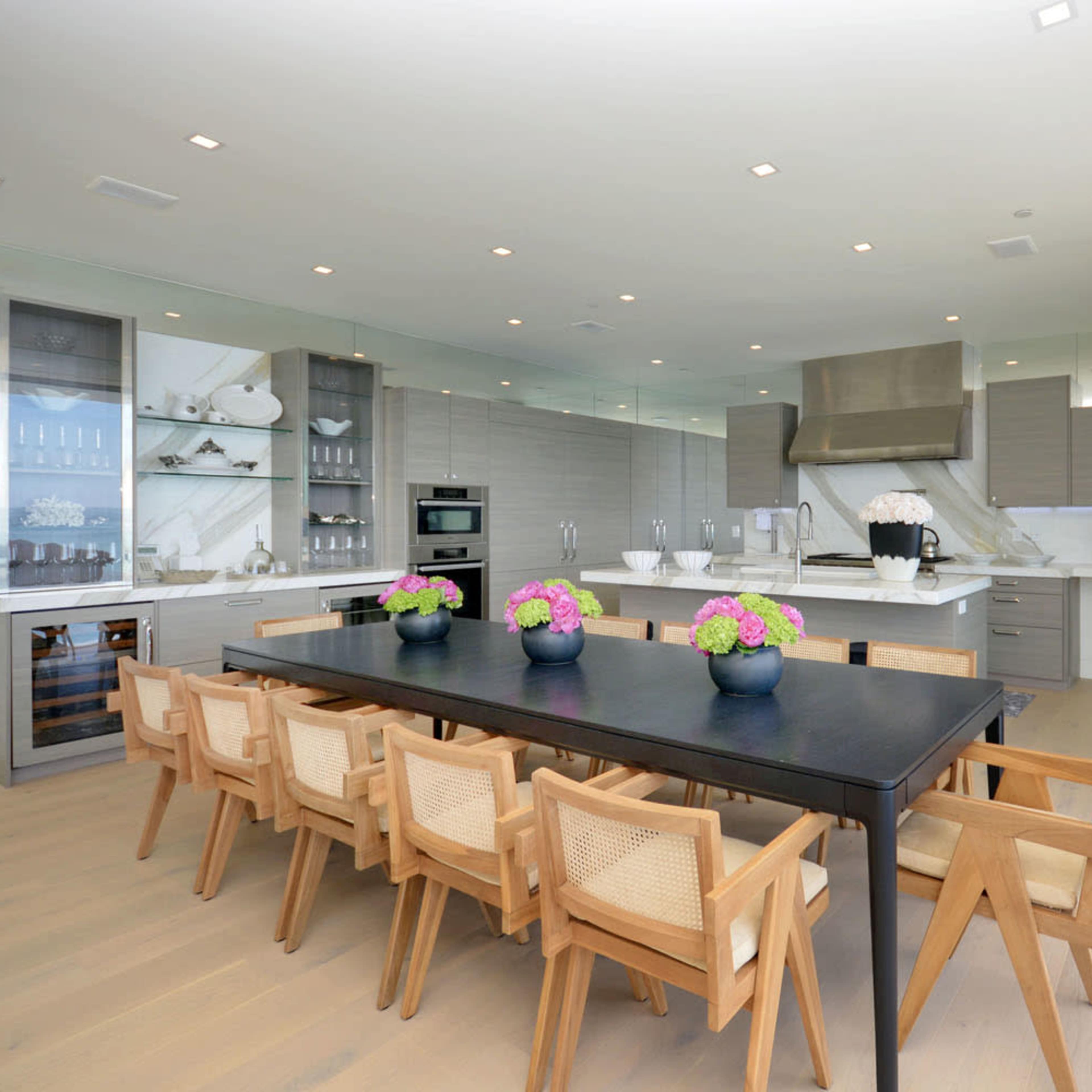 A modern kitchen and dining area features a large black table surrounded by wooden chairs, with a sleek kitchen equipped with stainless steel appliances and white cabinetry.