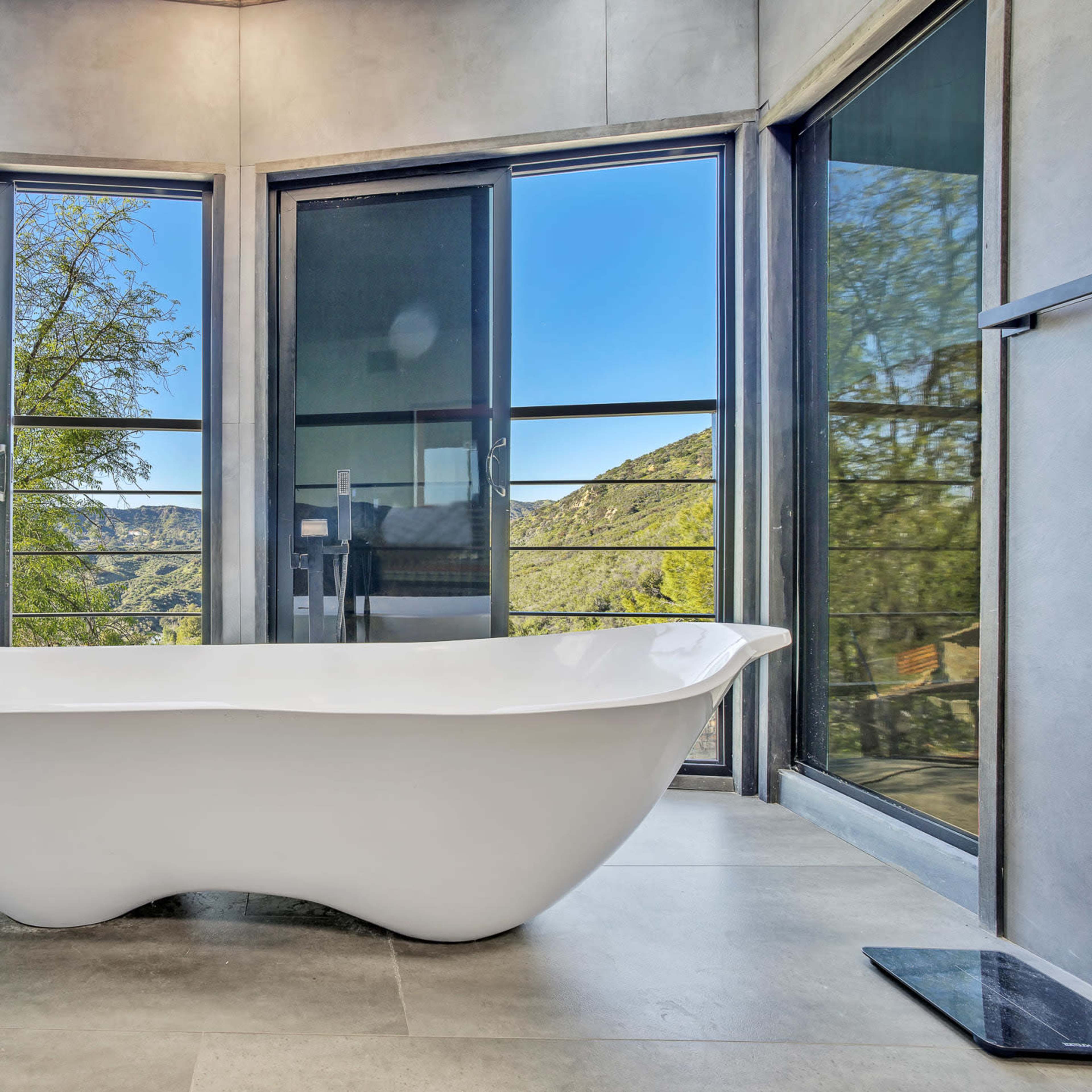 A freestanding white bathtub is centrally positioned in a modern bathroom with large windows that offer a view of a mountain landscape.