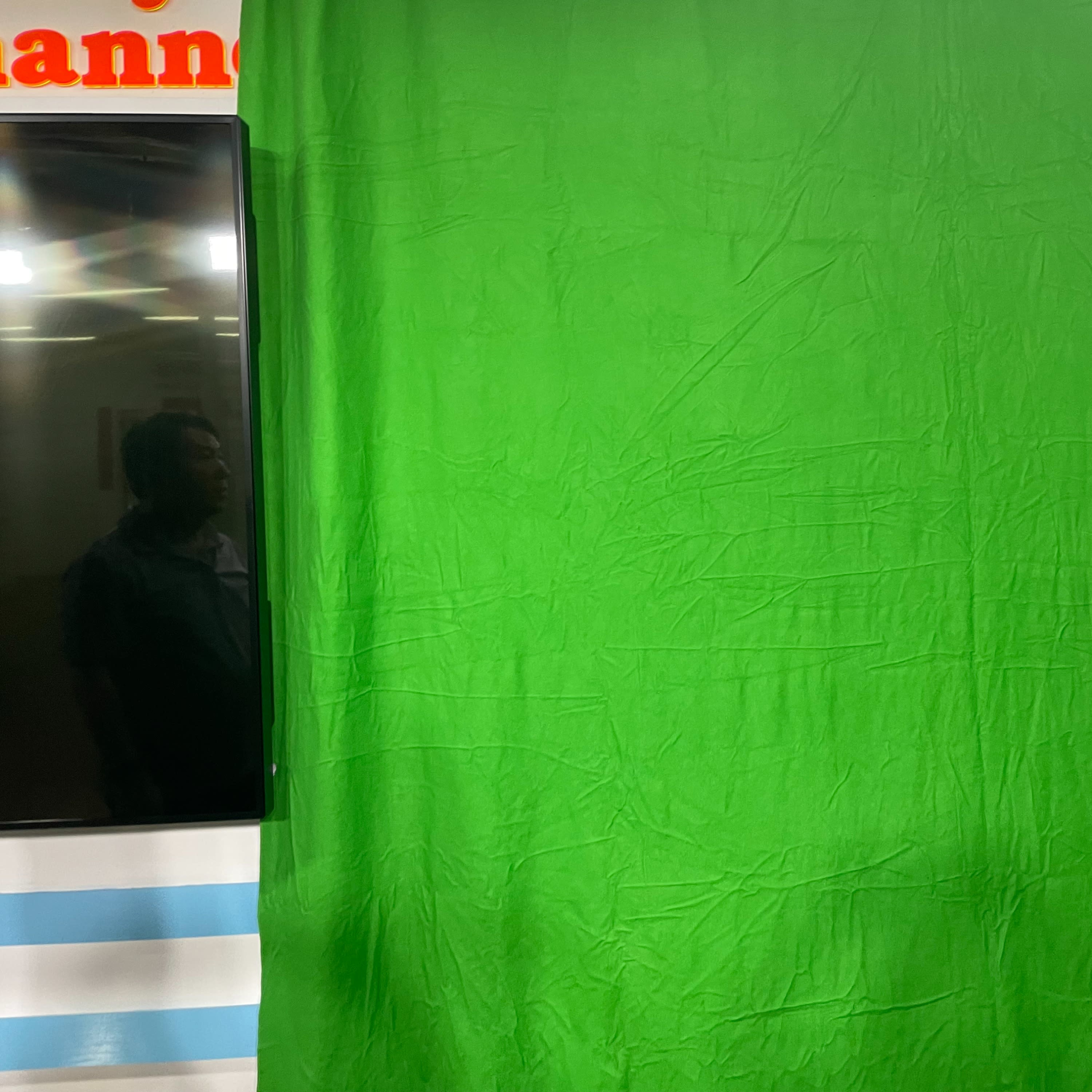 A green backdrop, commonly used for chroma keying, alongside a television screen and a person reflected in the glass.