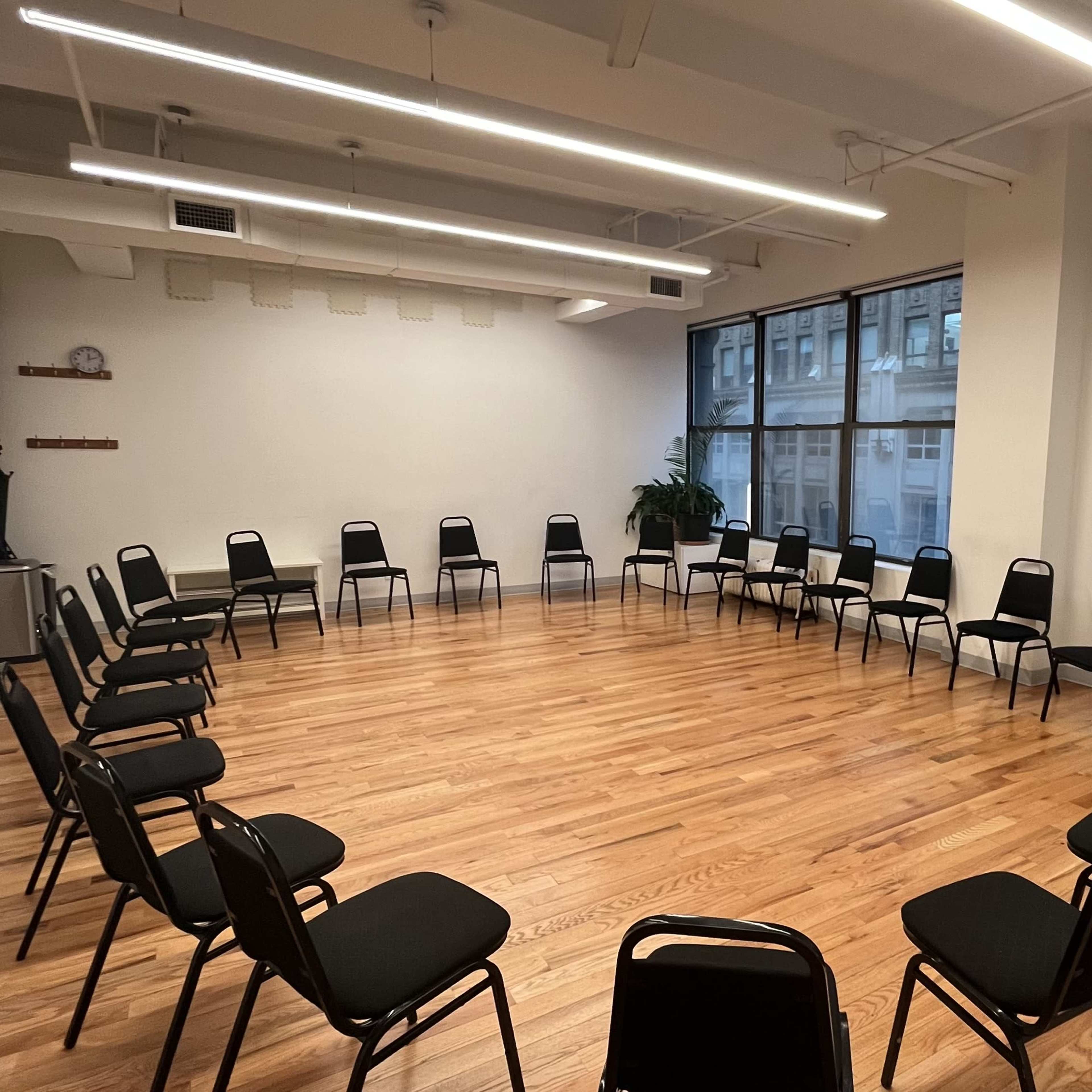 A spacious room with wooden floors contains two circular arrangements of black chairs facing each other, surrounded by large windows.