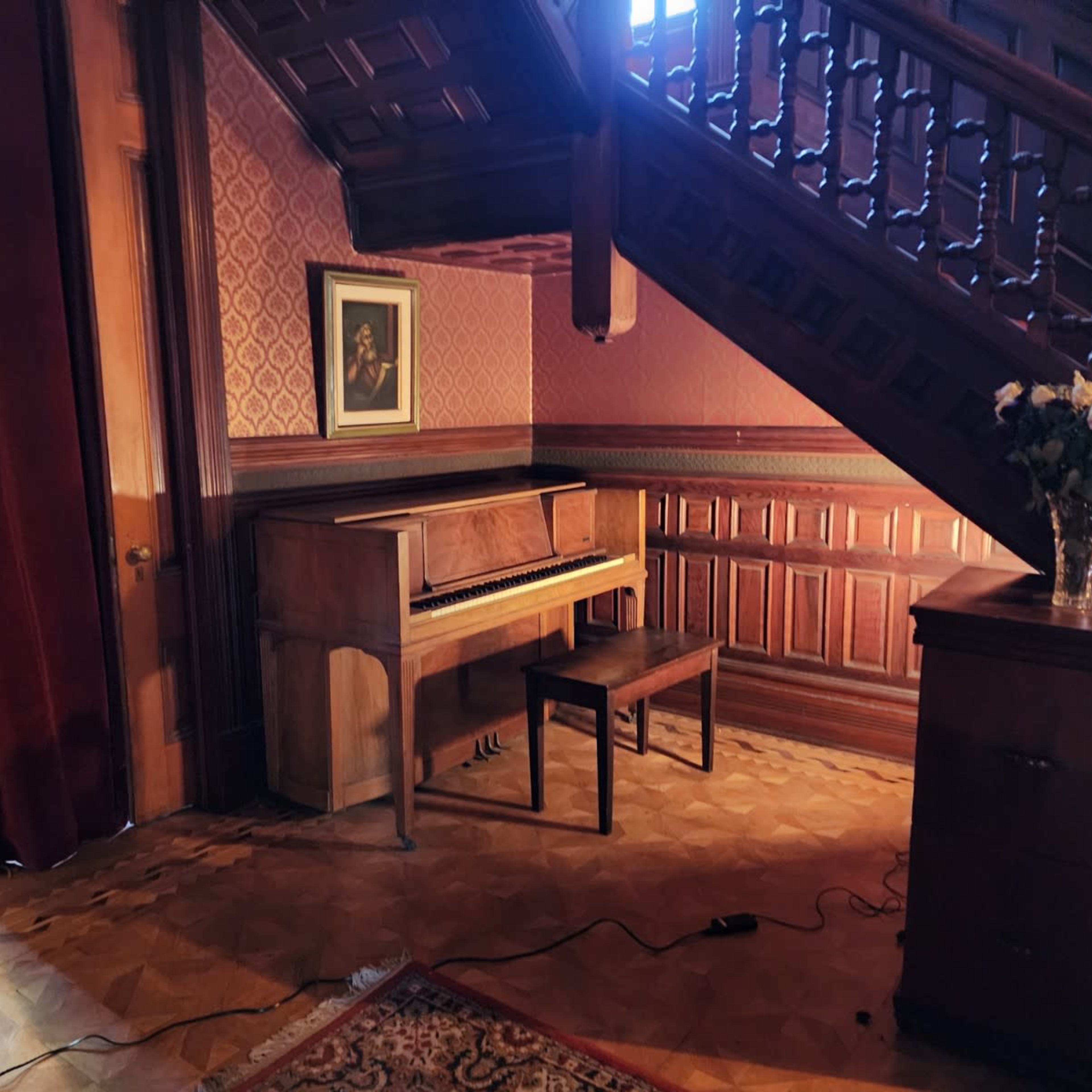 A wooden piano and bench are positioned under a staircase in a room with patterned wallpaper and a framed portrait on the wall.