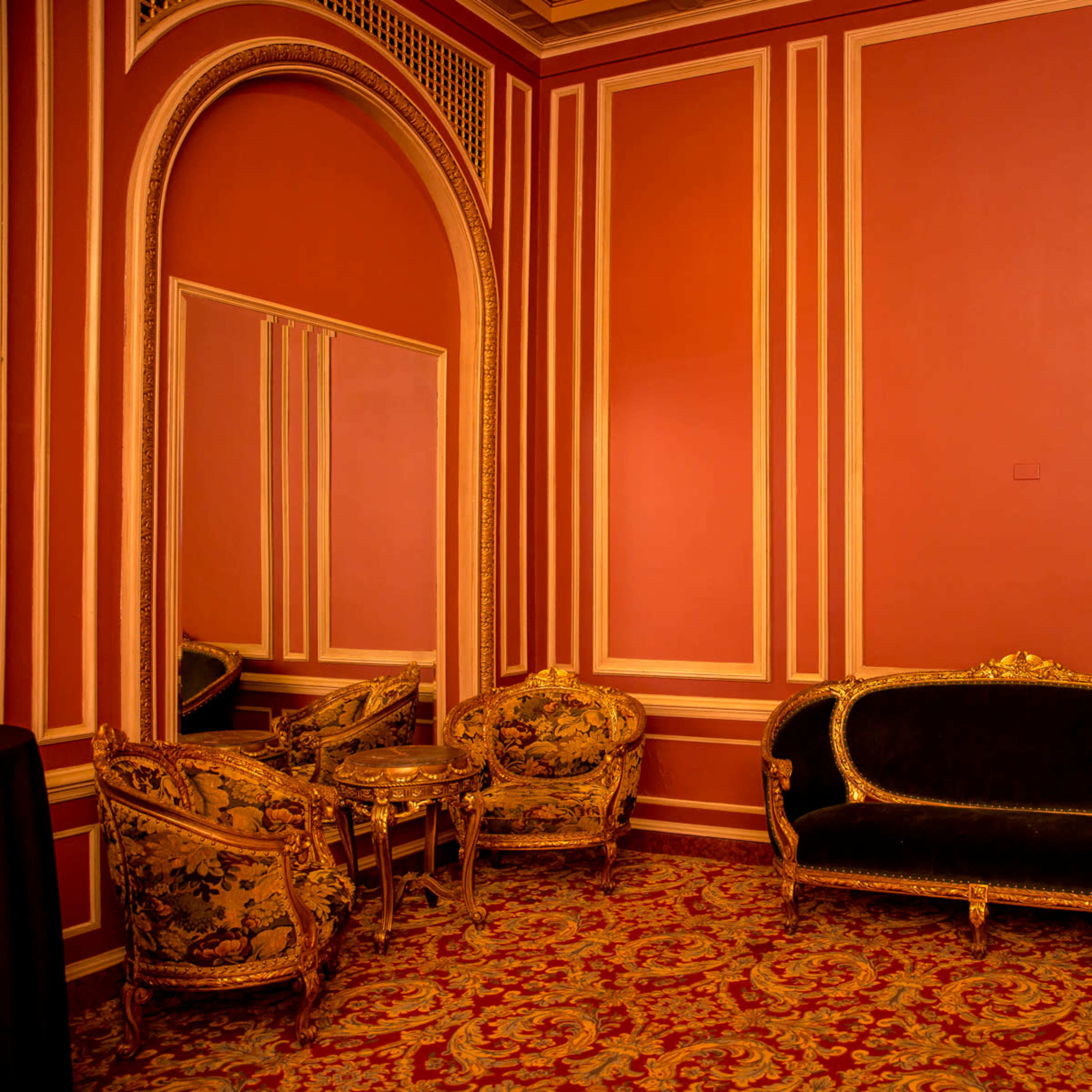 An ornate seating area with a black velvet sofa and floral-patterned armchairs against a richly colored wall, featuring a large mirror and decorative trim.