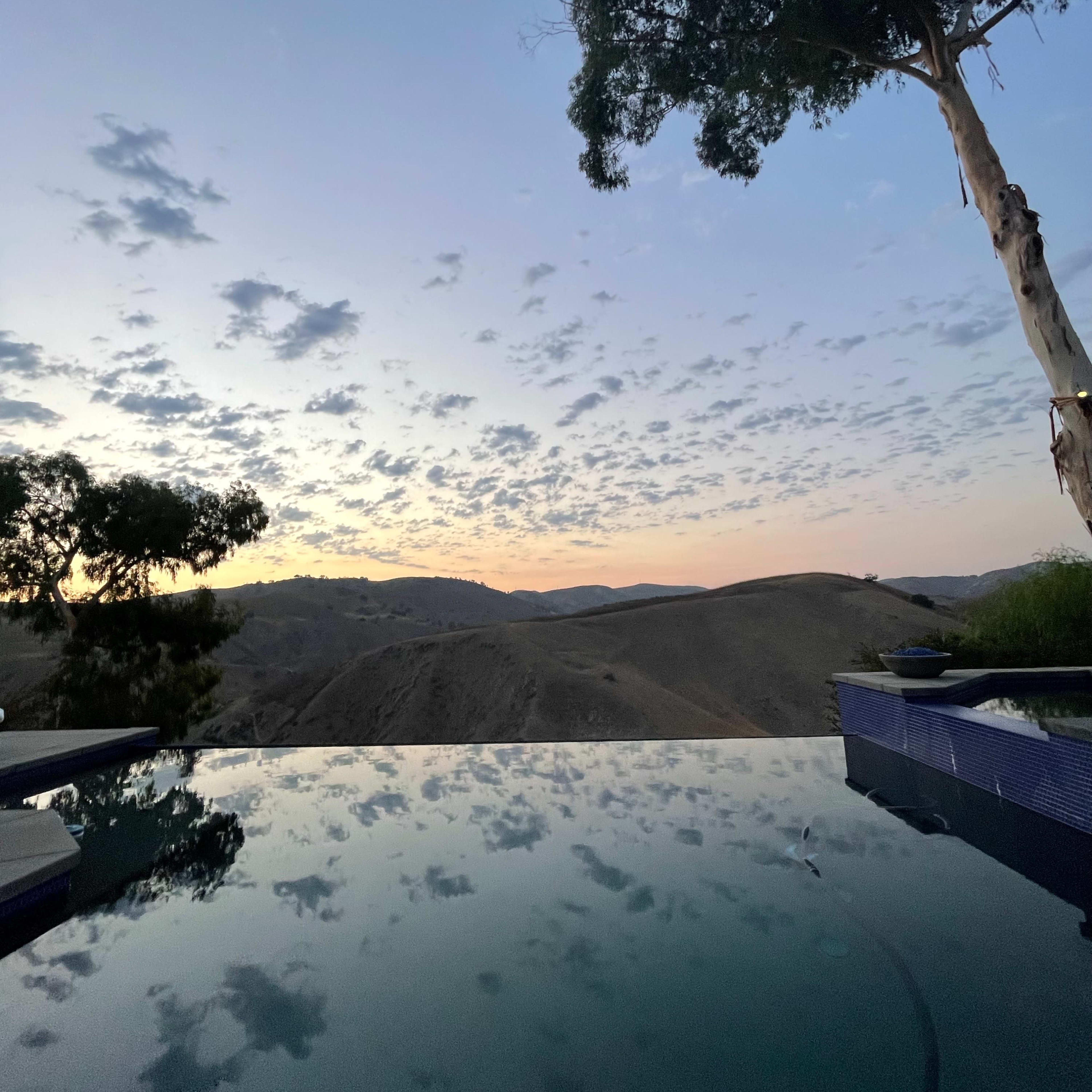 An infinity pool reflecting the sky and surrounding hills at dusk.