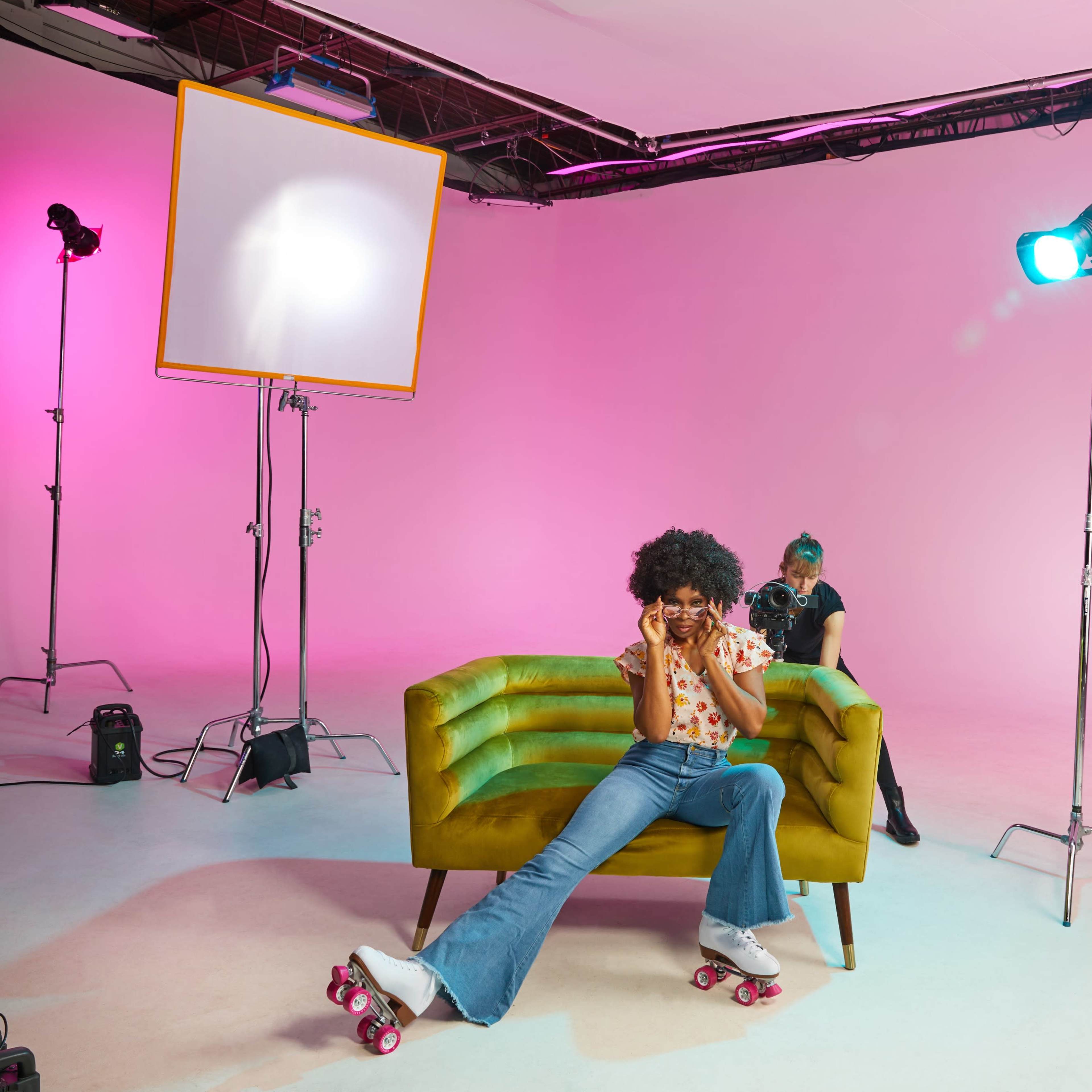 A person wearing roller skates sits on a green couch in a studio with pink walls and multiple lights set up for a photoshoot.