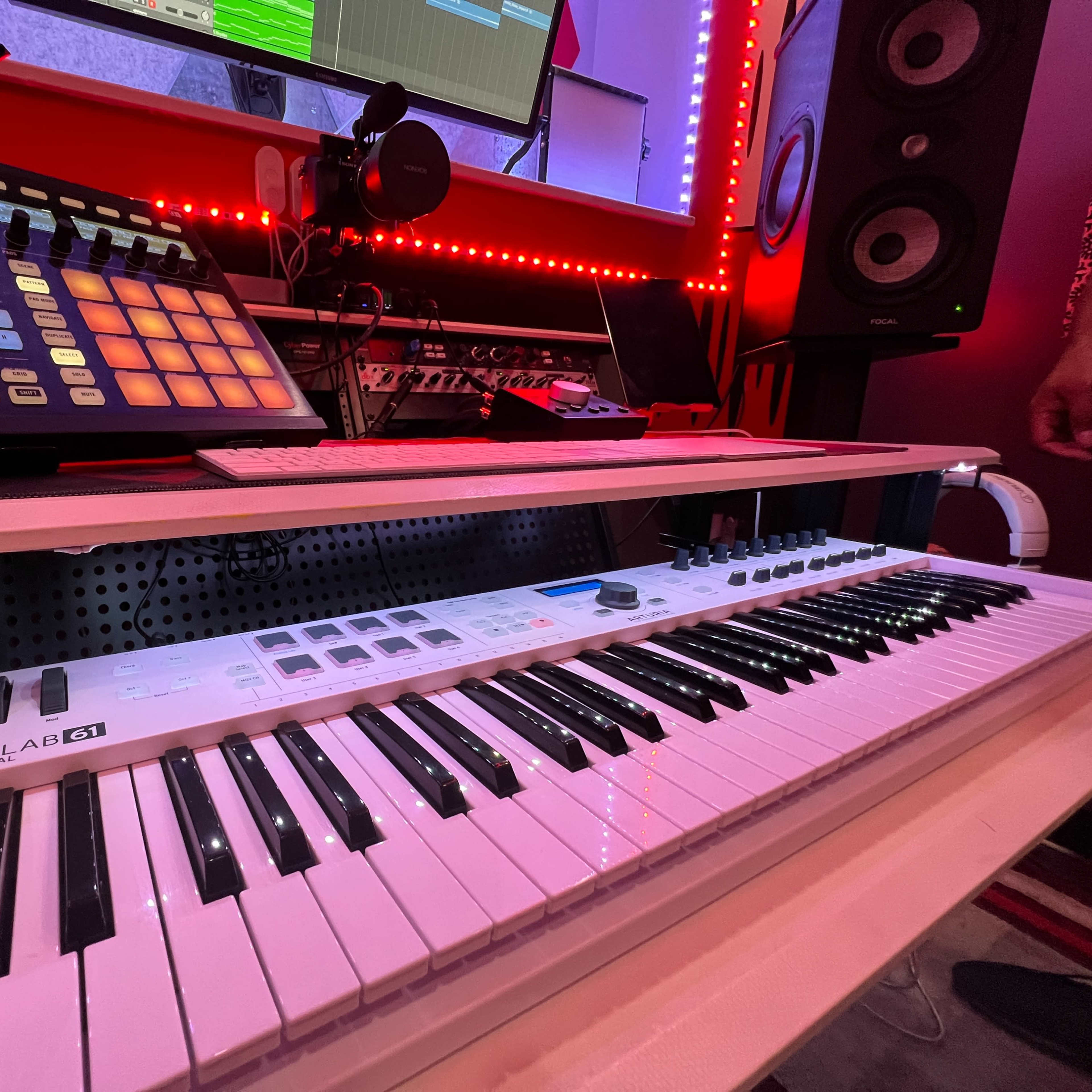 A music production setup featuring a MIDI keyboard and a drum pad controller, along with studio monitors and computer screens in the background.