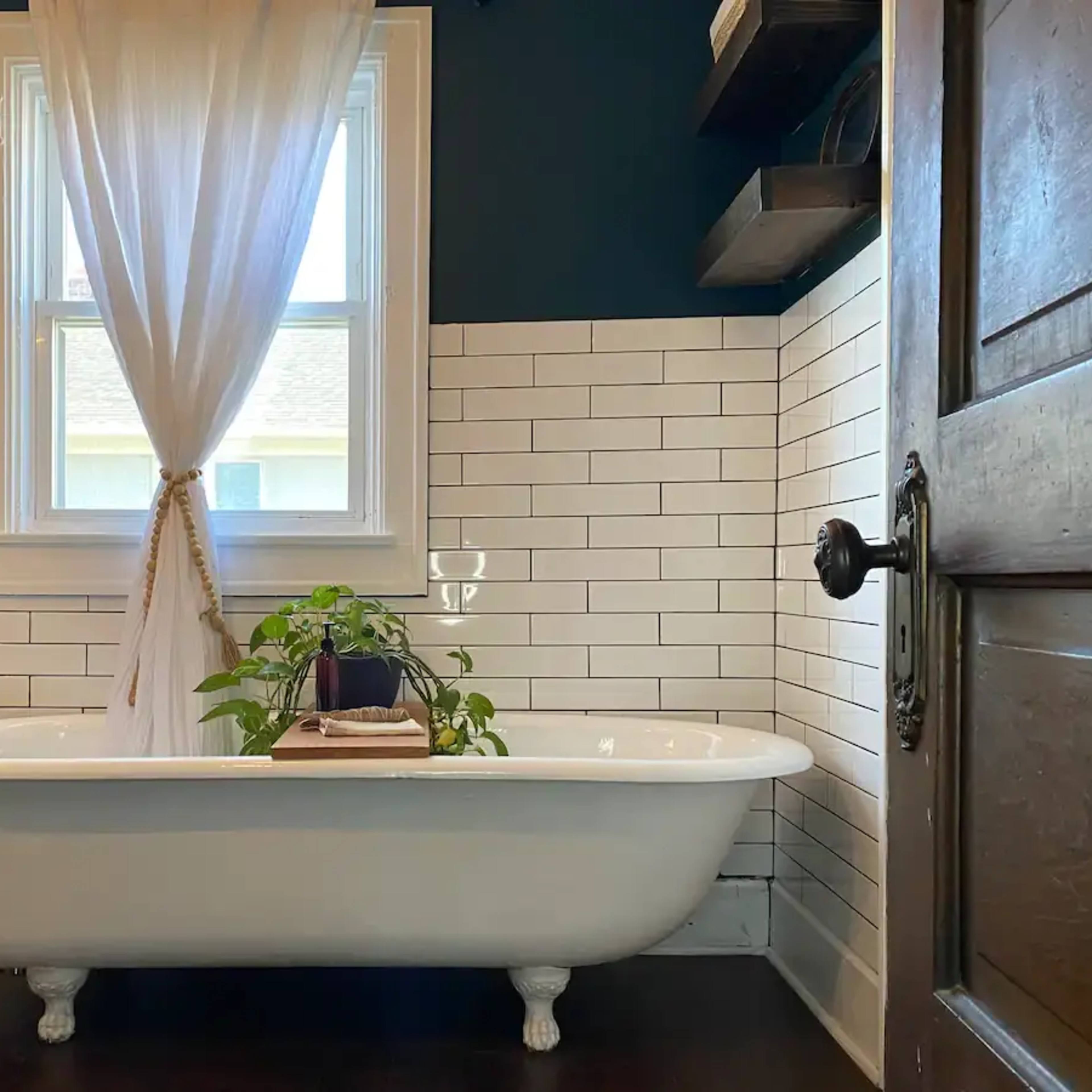 A clawfoot bathtub is positioned in a bathroom with white subway tile walls and a dark wooden door, featuring a plant and a glass of water on a wooden tray beside it.