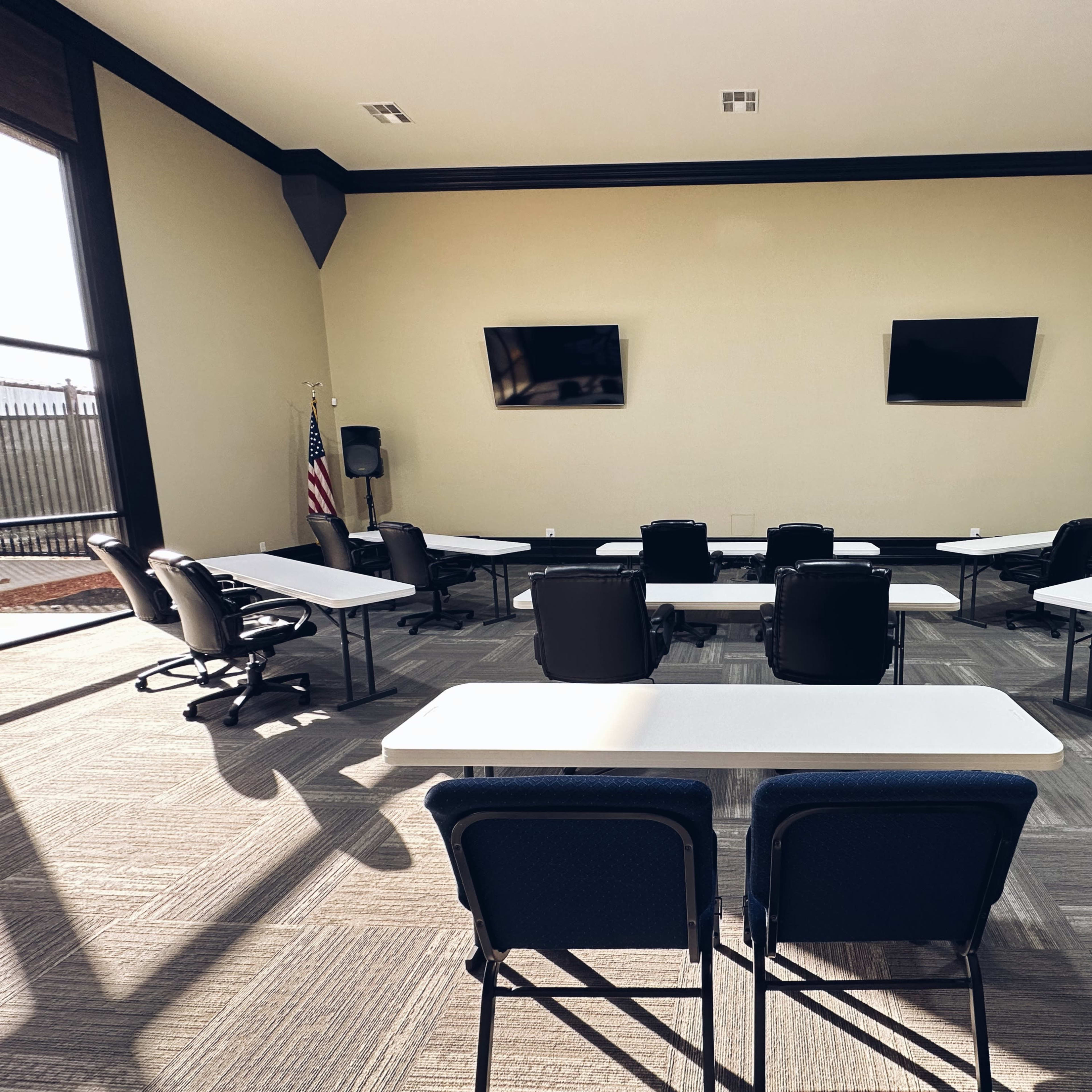 A conference room features several tables and chairs arranged in a U-shape, with two televisions mounted on the wall and windows allowing natural light.