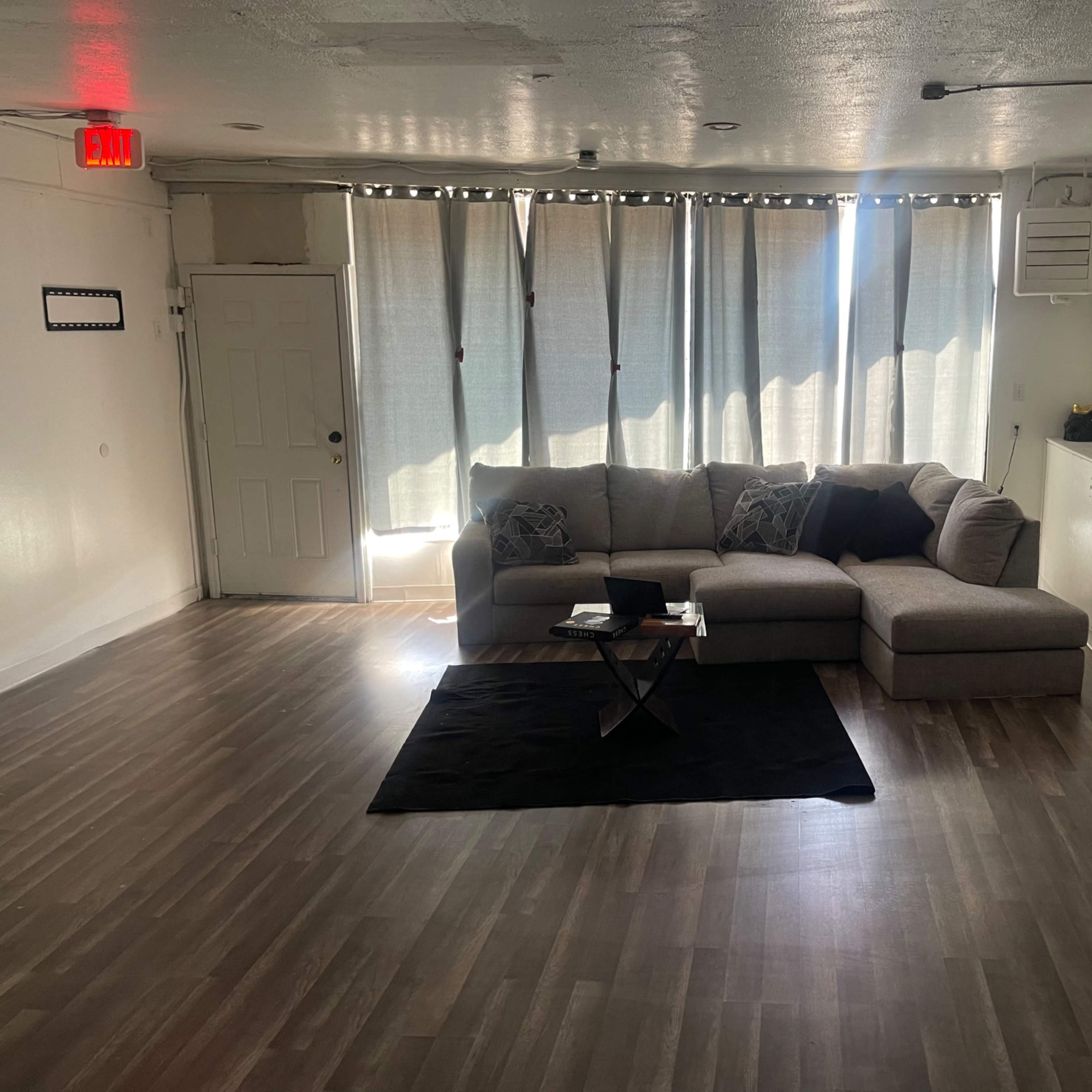 A spacious, minimally furnished living room featuring a large sectional sofa, a coffee table on a black rug, and natural light streaming through sheer curtains.