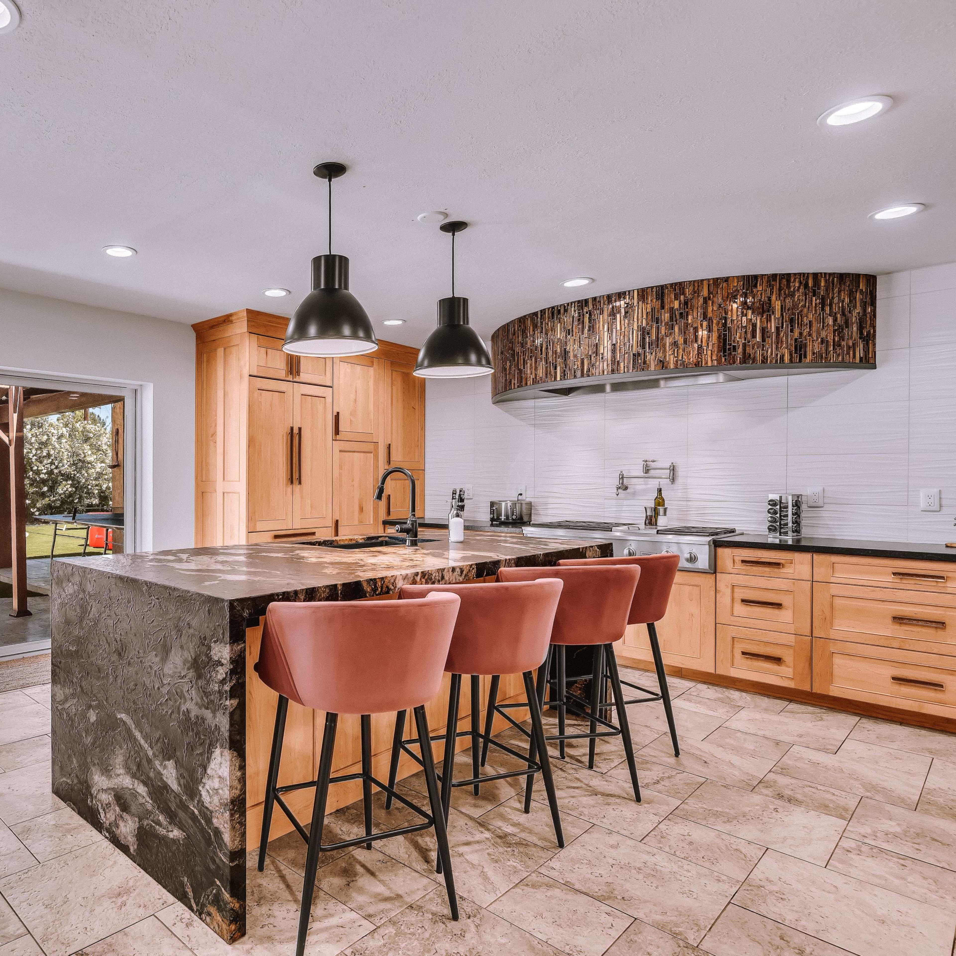 A modern kitchen featuring a large island with bar stools, light wood cabinetry, and a view of an outdoor patio through sliding glass doors.