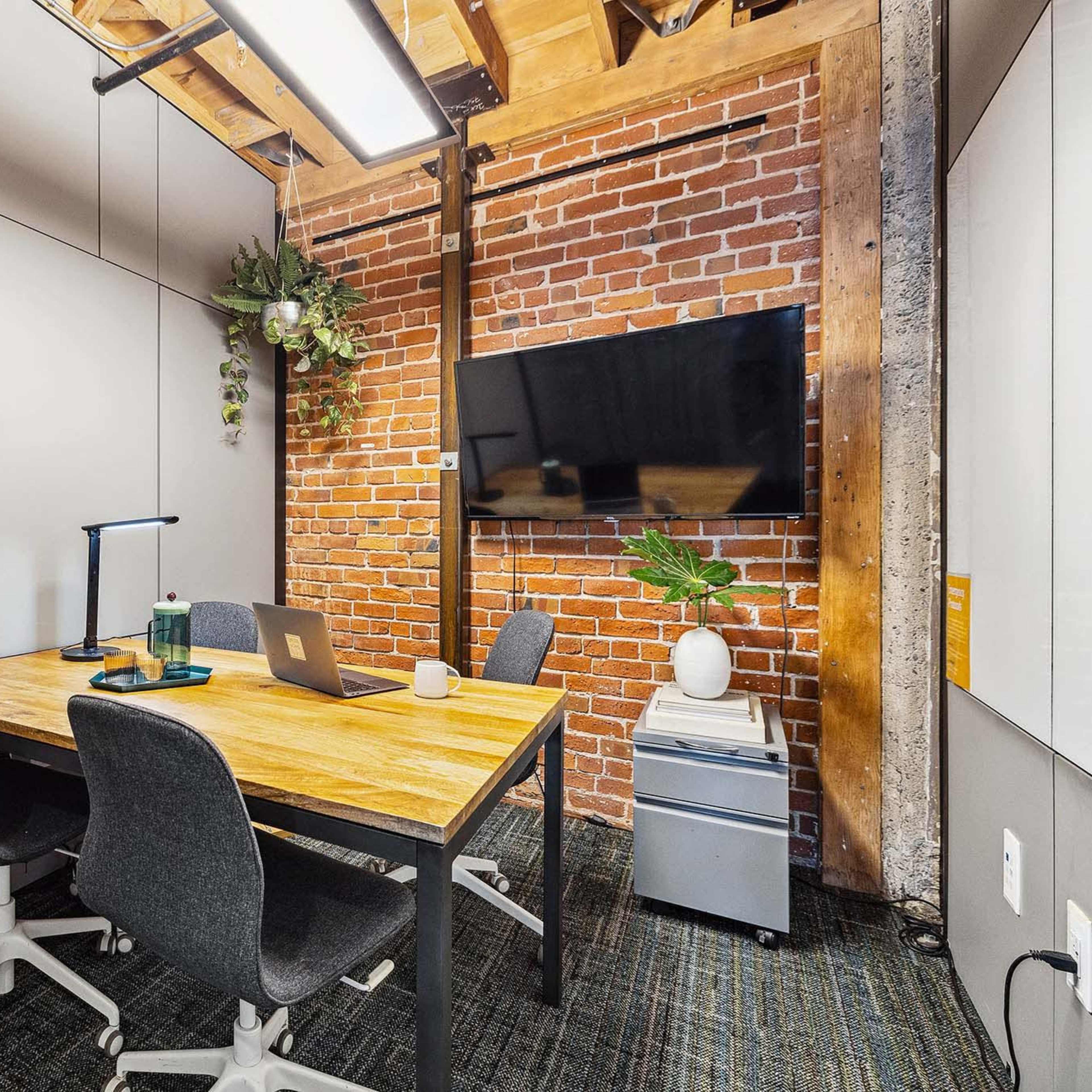 A small meeting room features a wooden table, three chairs, a large wall-mounted TV, and exposed brick walls with greenery decor.