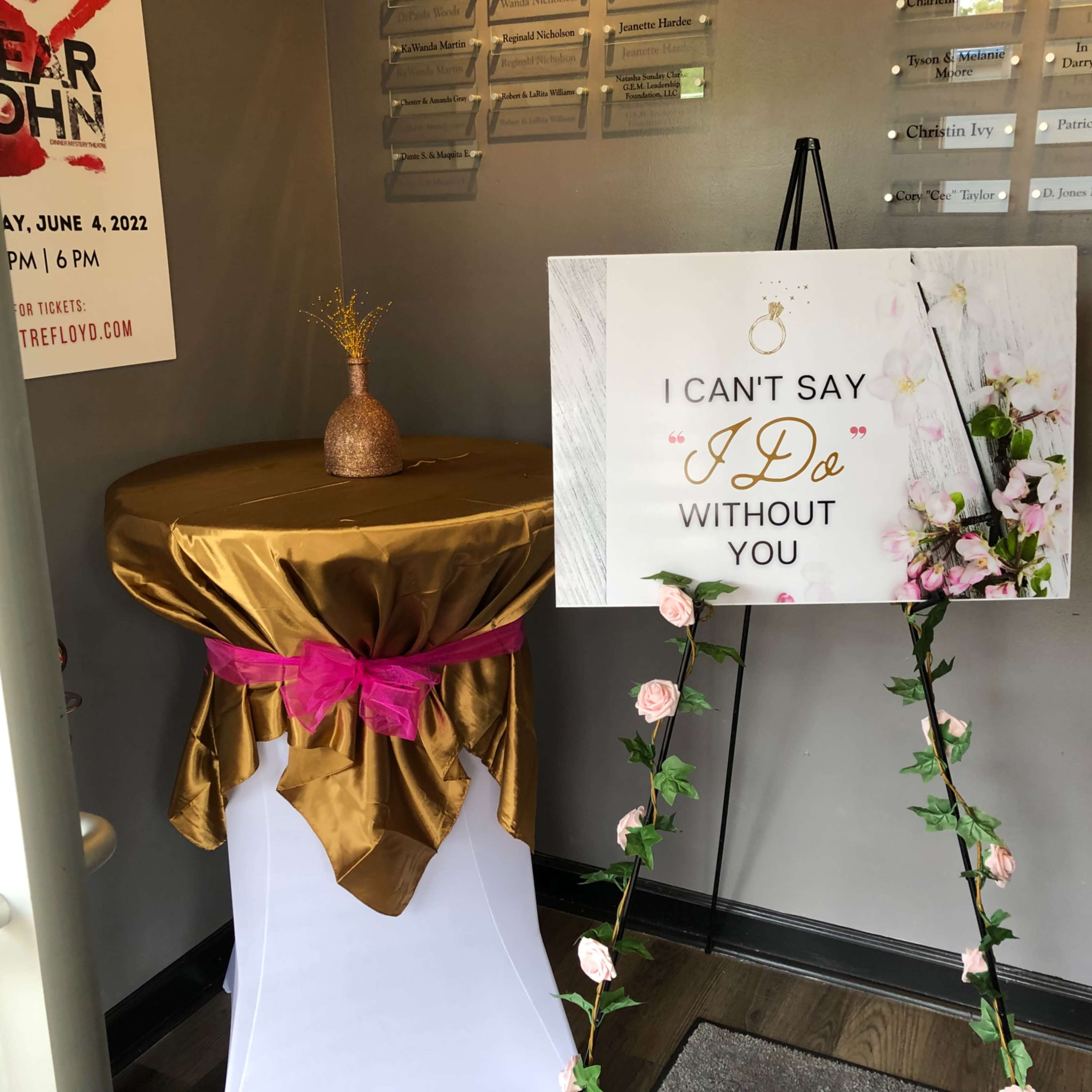 A decorated area with a gold tablecloth, a sign that reads "I can't say 'I do' without you," and floral accents.