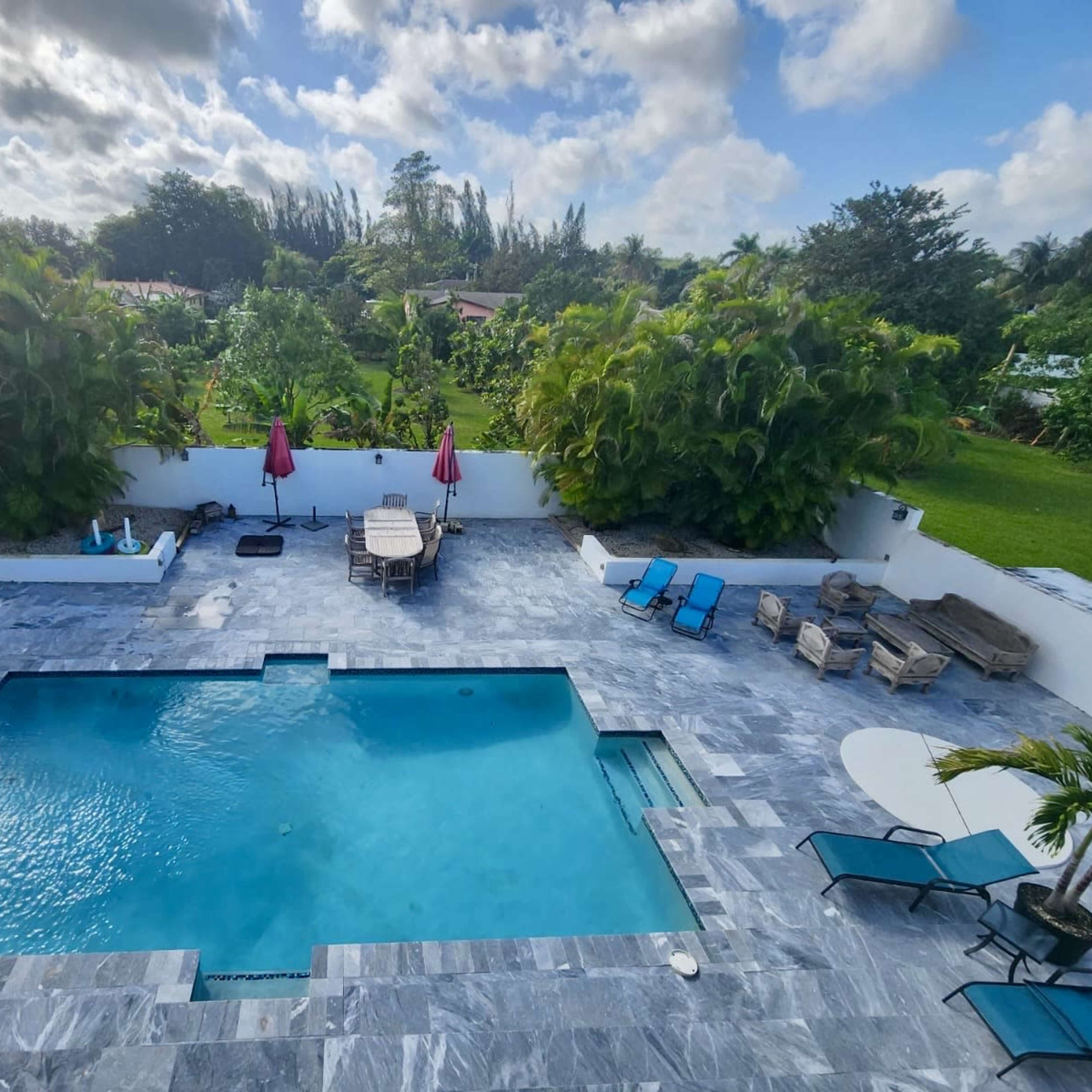 A large rectangular swimming pool surrounded by lounge chairs and tables, set in a landscaped backyard with lush greenery and palm trees.
