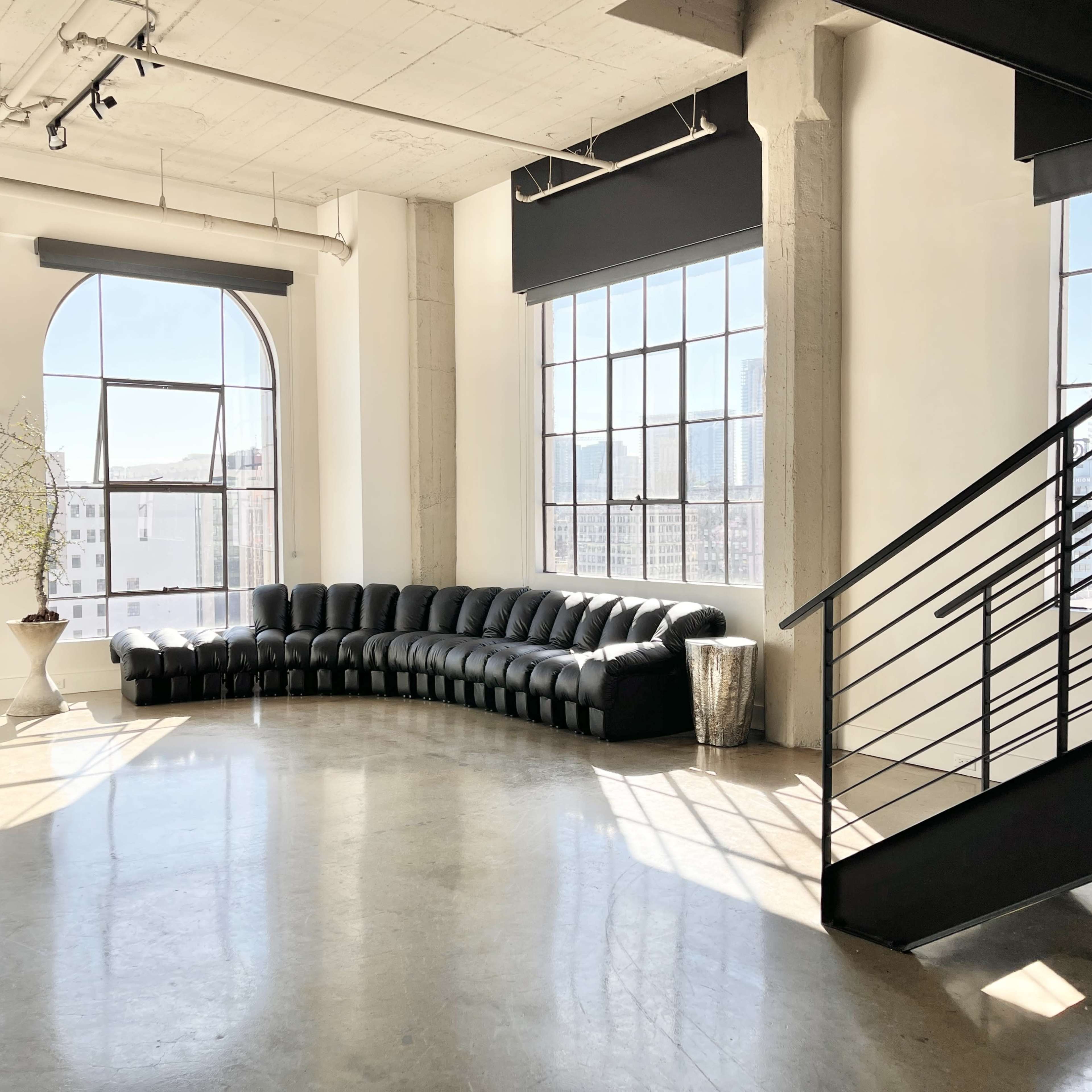 A spacious room with large arched windows, featuring a black curved sofa and a metal side table, illuminated by natural light.