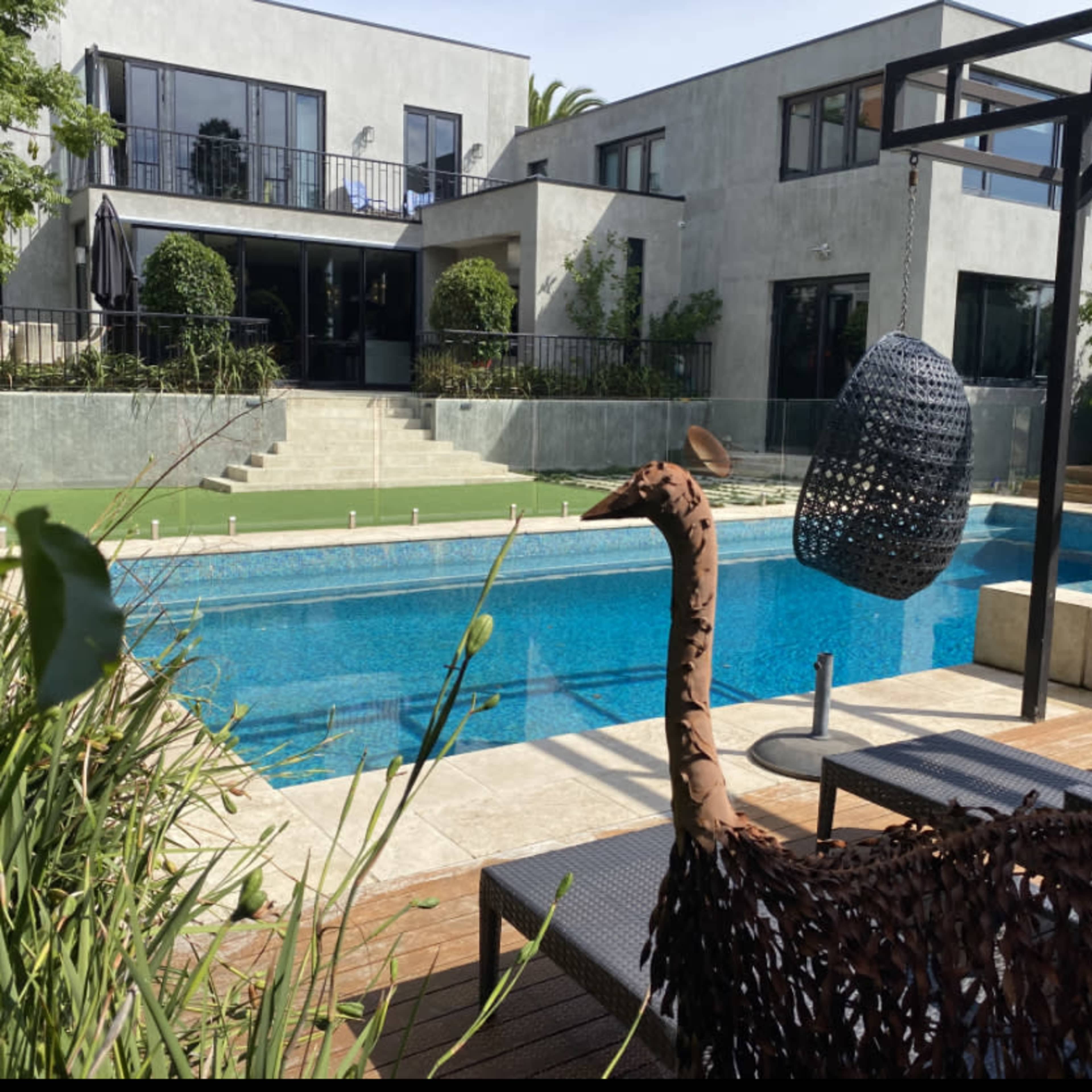 A metal sculpture of an ostrich stands near a swimming pool, with a modern house and landscaped greenery in the background.