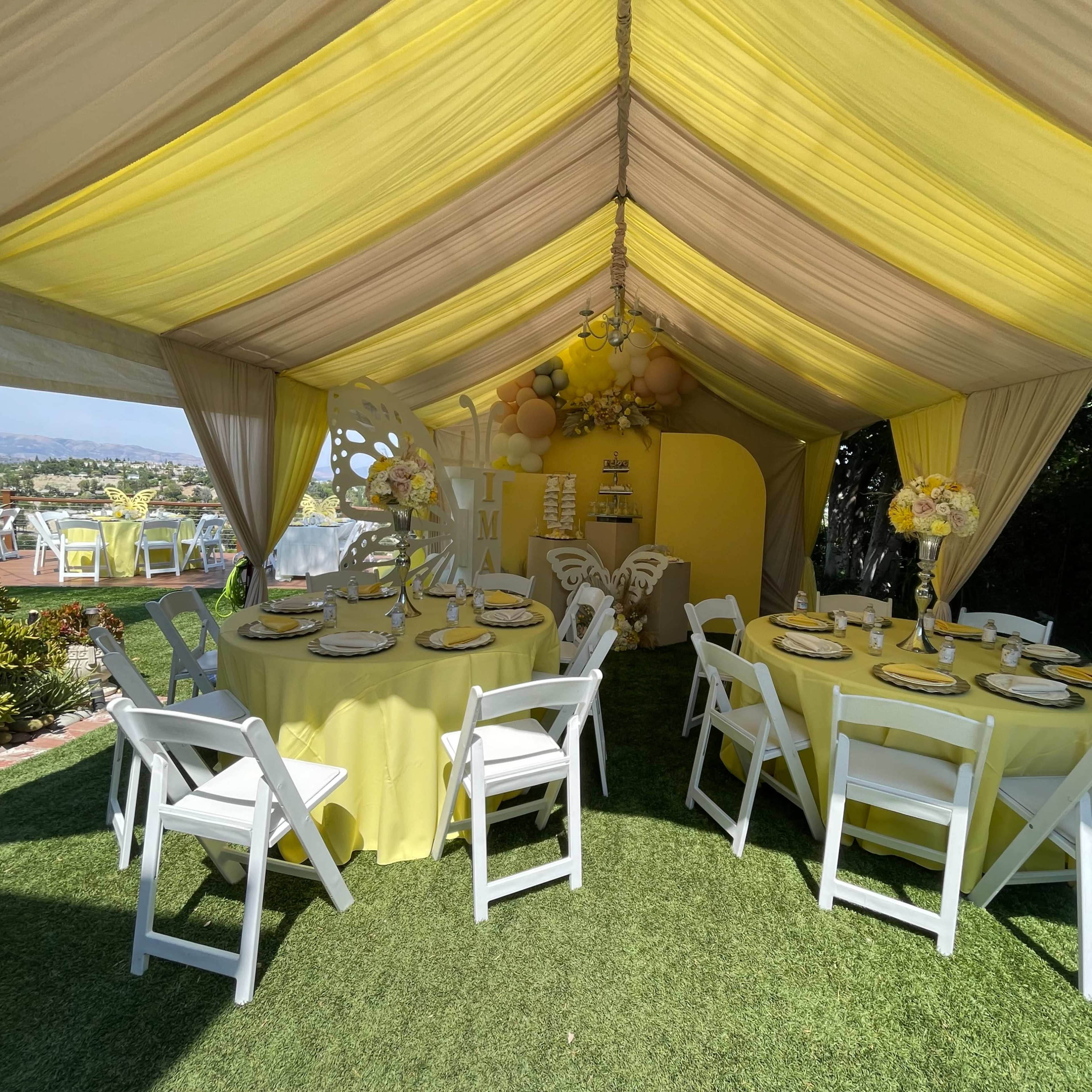 A tented dining area with yellow decorations features tables set with white chairs and a backdrop adorned with balloons and floral arrangements.