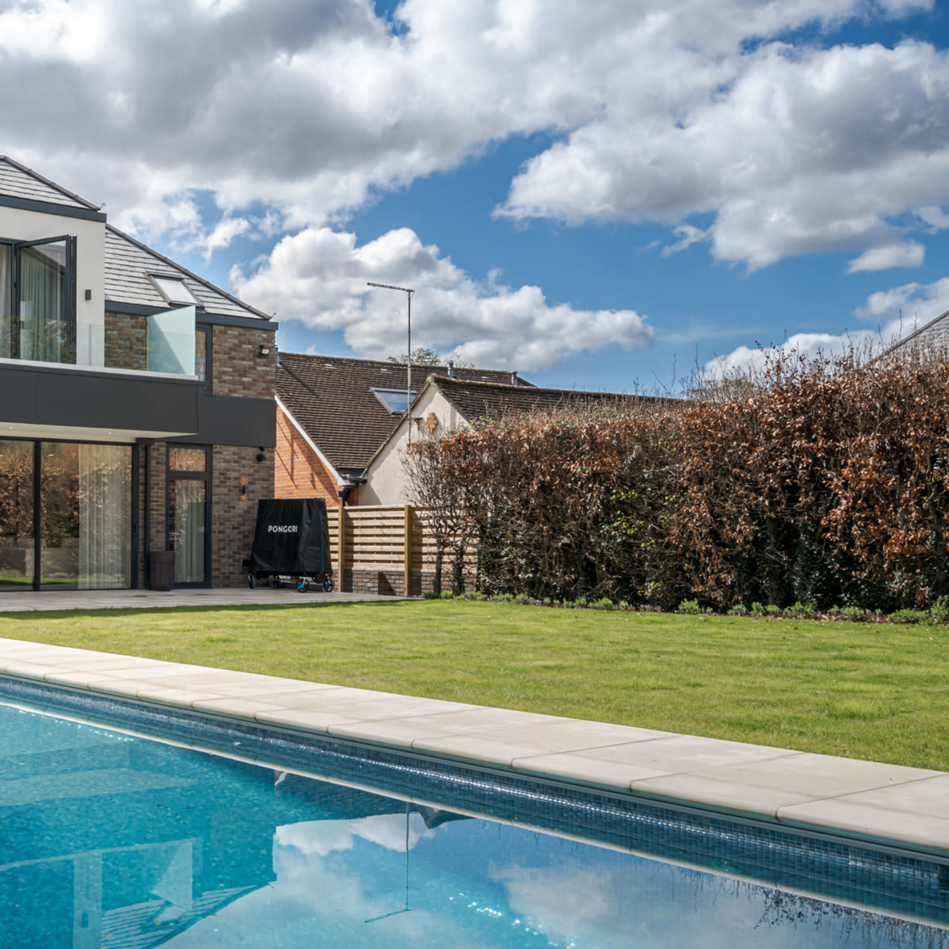 A modern two-story house with large windows overlooks a swimming pool and green lawn under a partly cloudy sky.
