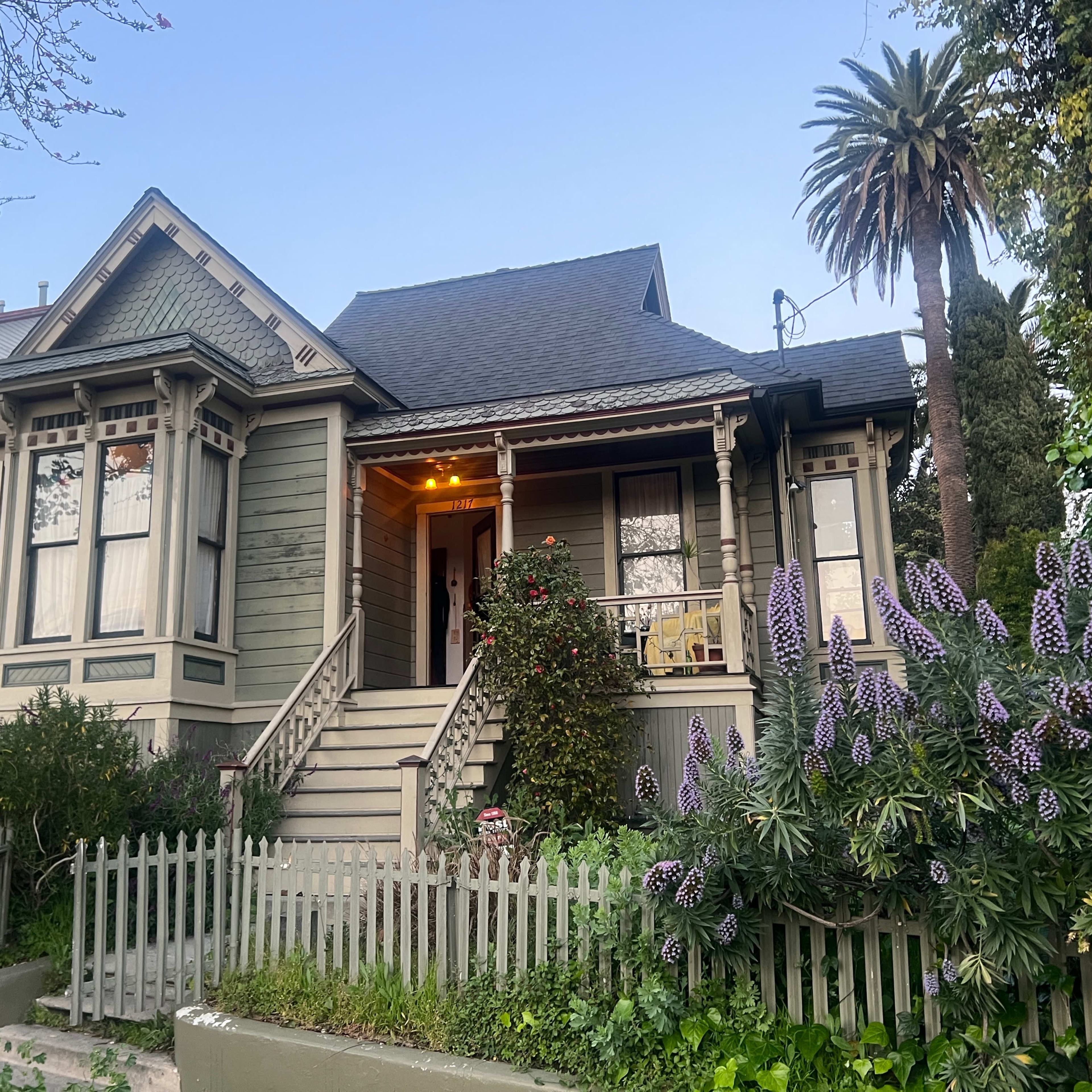 A two-story Victorian-style house with a front porch, surrounded by green foliage and blooming flowers, is located on a well-maintained property.