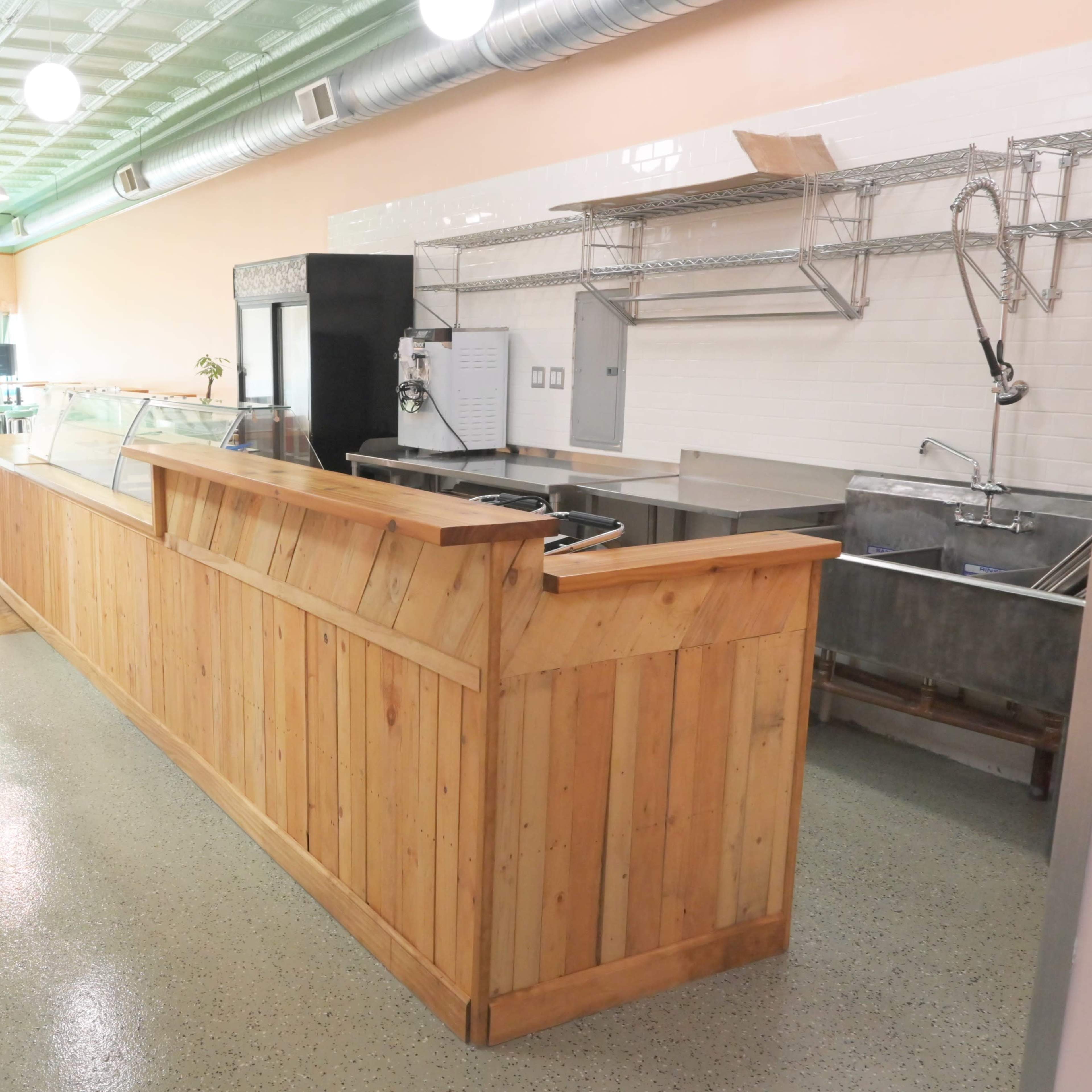 A spacious, empty café interior featuring a wooden counter with glass display cases, stainless steel sinks, and a tiled wall.